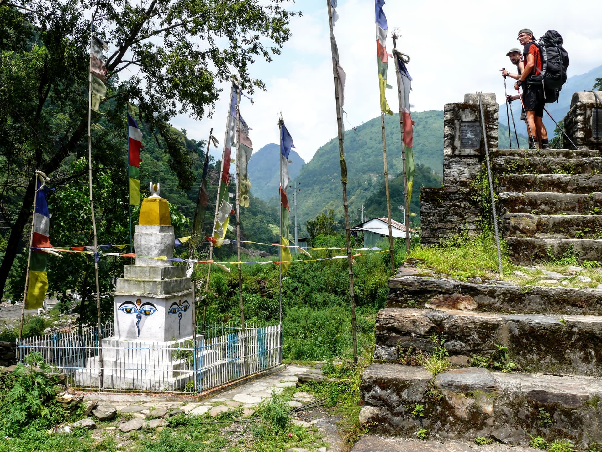 Hikers with backpacks stand near prayer flags and a stupa in lush green mountains.