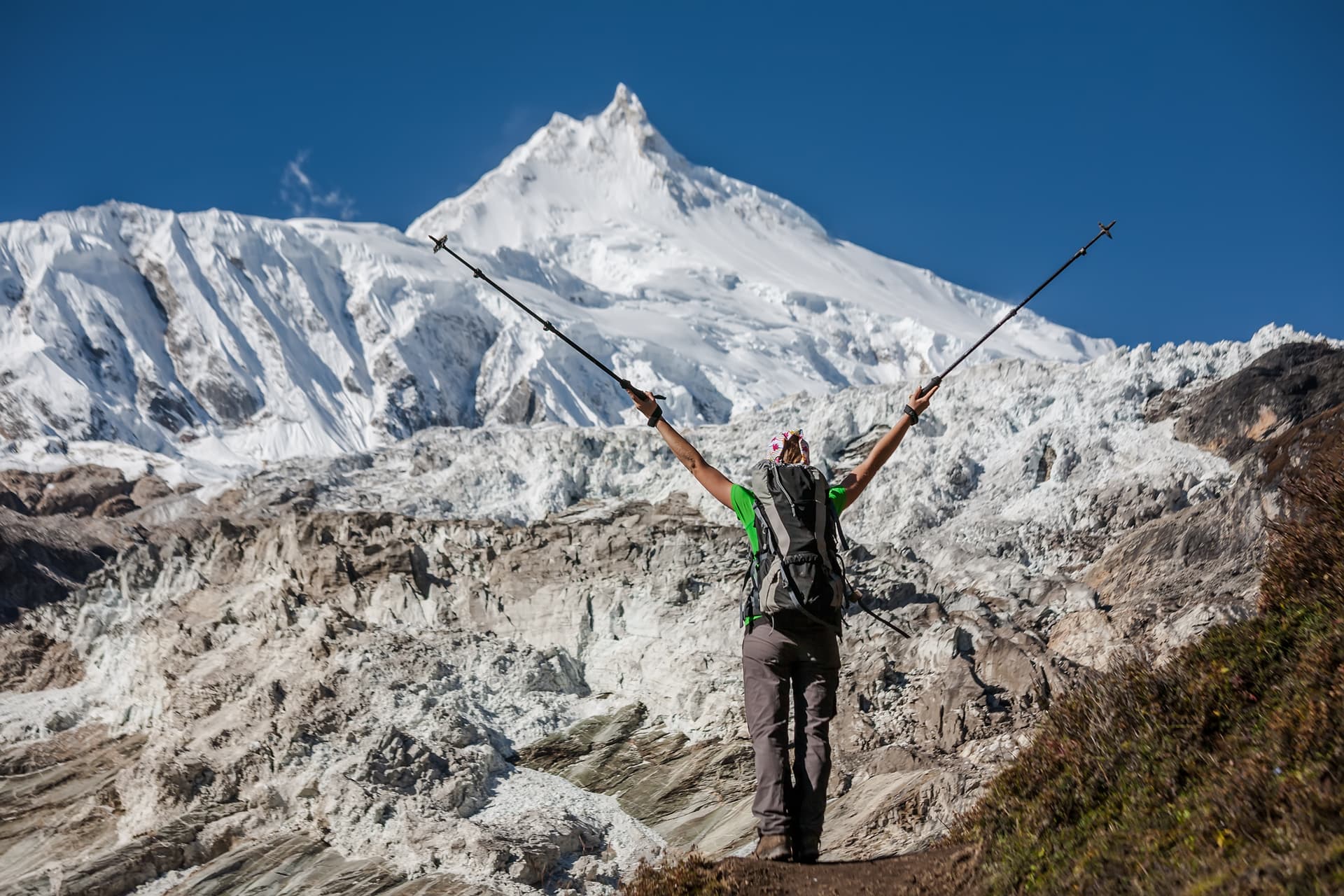 Hiker celebrating with trekking poles in front of snow-covered Manaslu mountain and glacier.