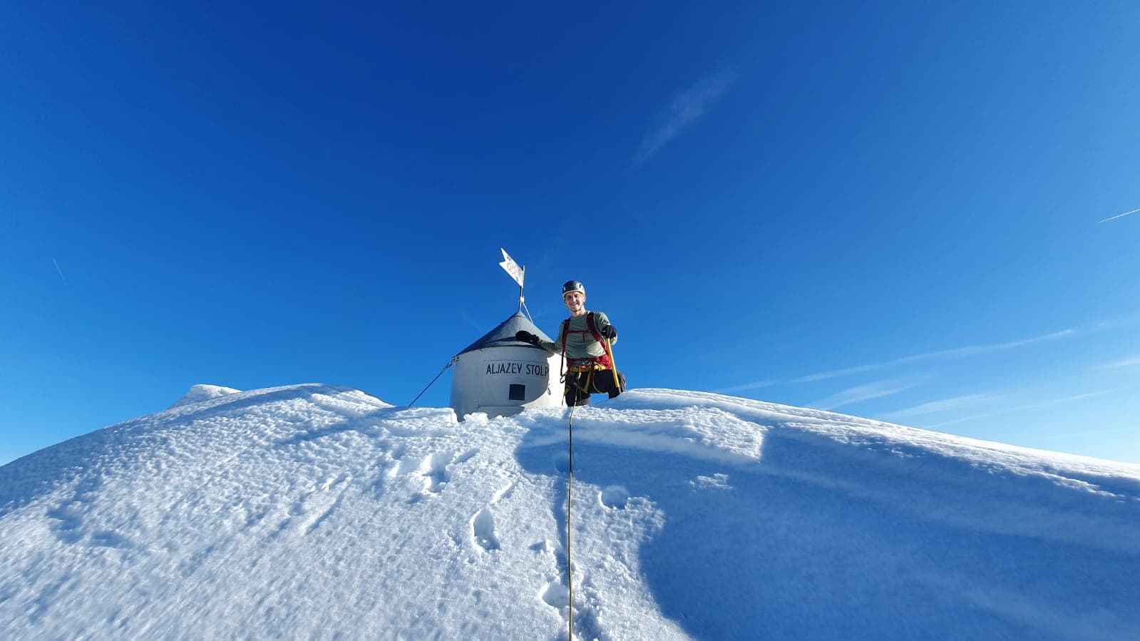 Hiker at Aljažev Stolb summit on Triglav mountain in deep winter snow under blue sky.