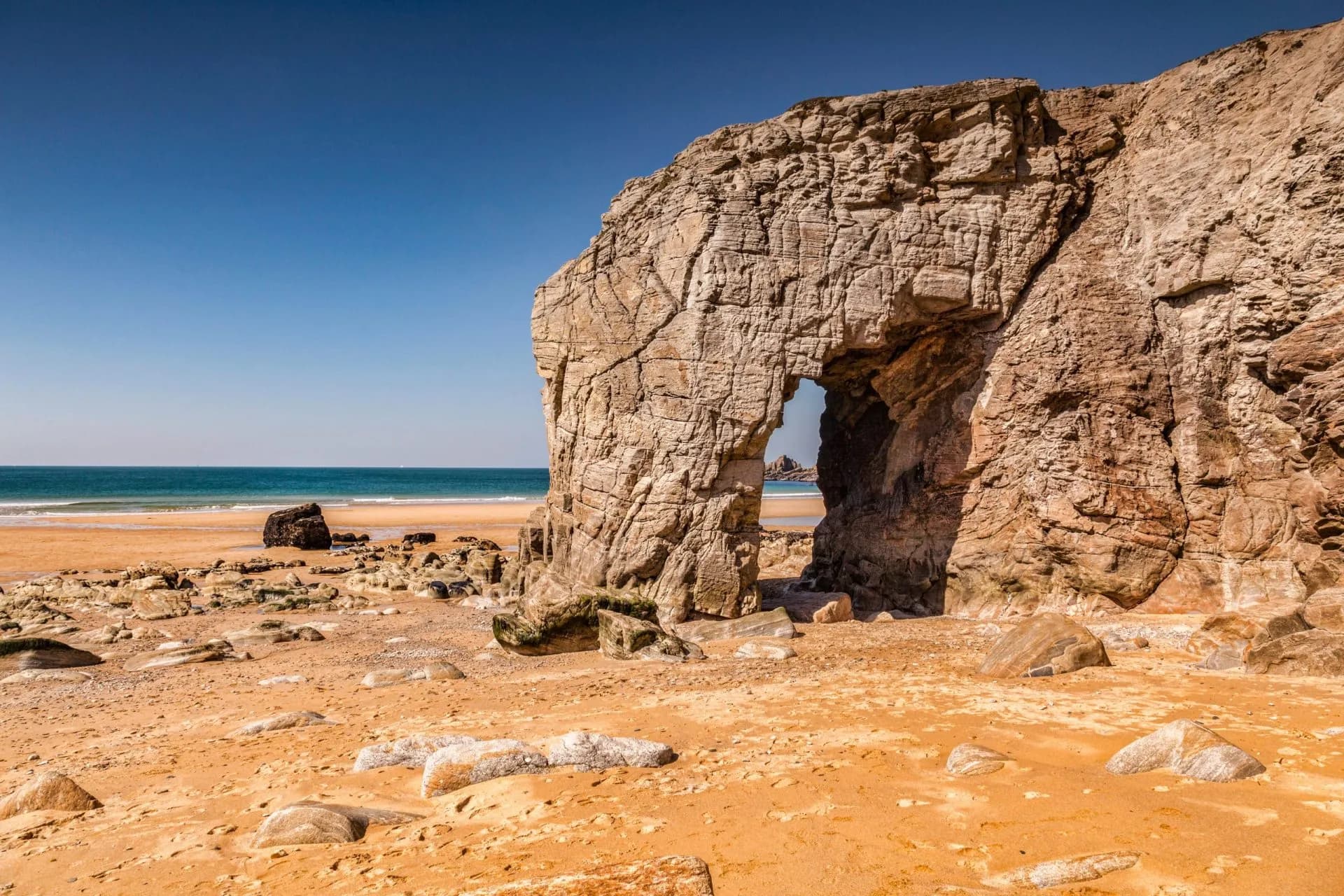Natural rock arch on sandy beach with boulders and blue ocean under clear sky at Port Blanc.