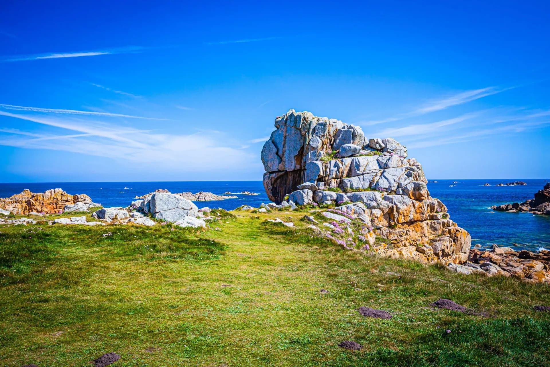 Large granite rock formation on grassy coastline overlooking deep blue sea under bright sky, Le Gouffre Coast.