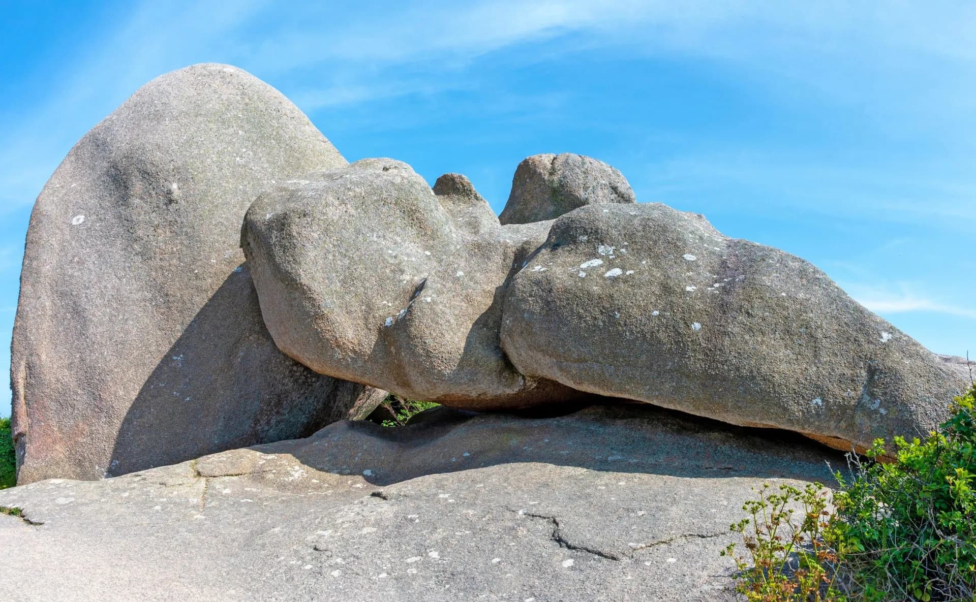 Large, rounded pink granite boulders stacked under a bright blue sky, with green scrub in the foreground.