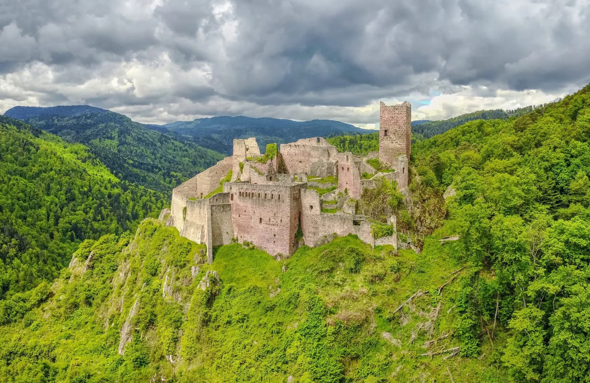 Ruins of Saint Ulrich Castle perched on a lush green, forested hillside under a cloudy sky.