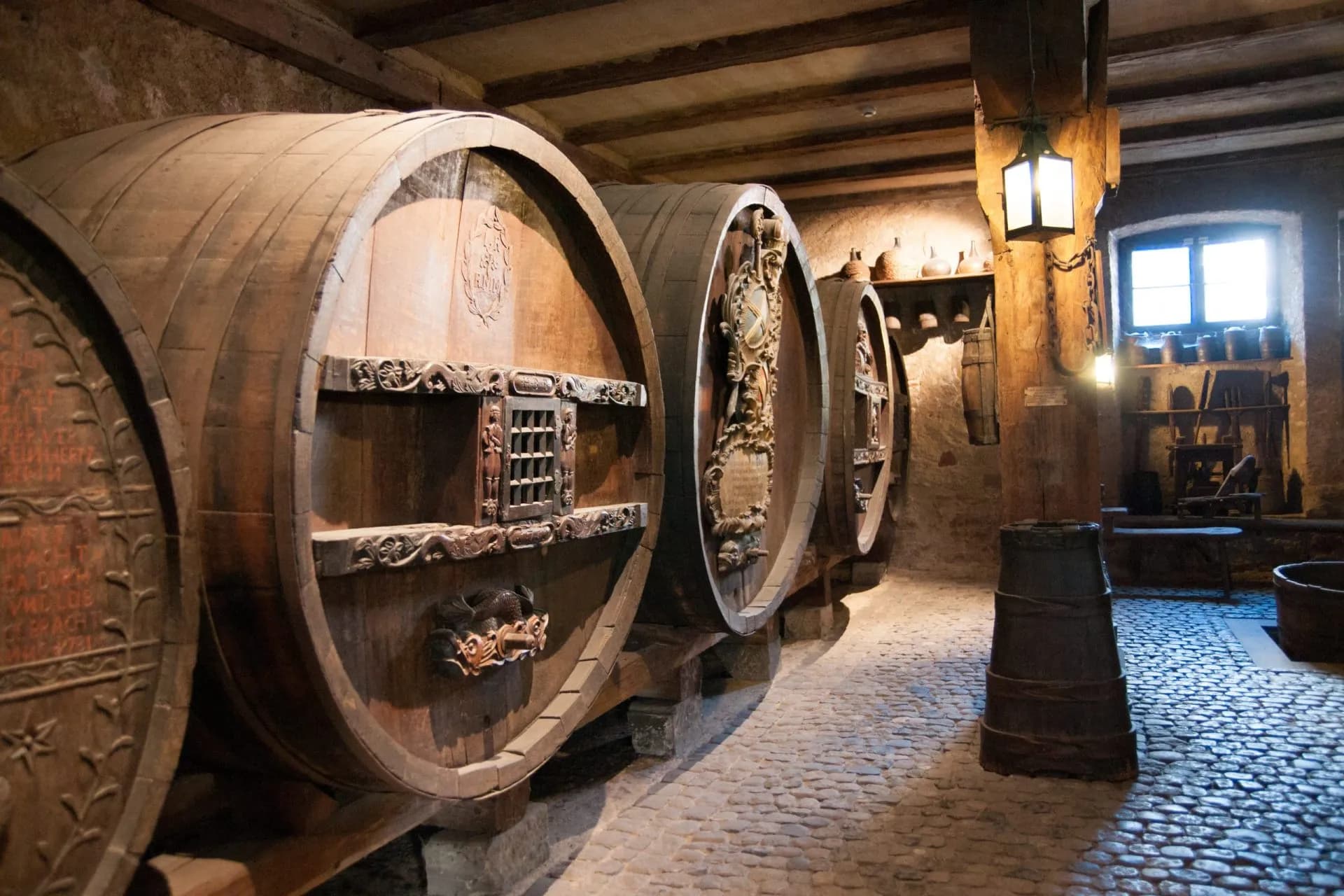 Large, ornate wooden wine barrels in a dimly lit cellar with a cobblestone floor, Alsace.
