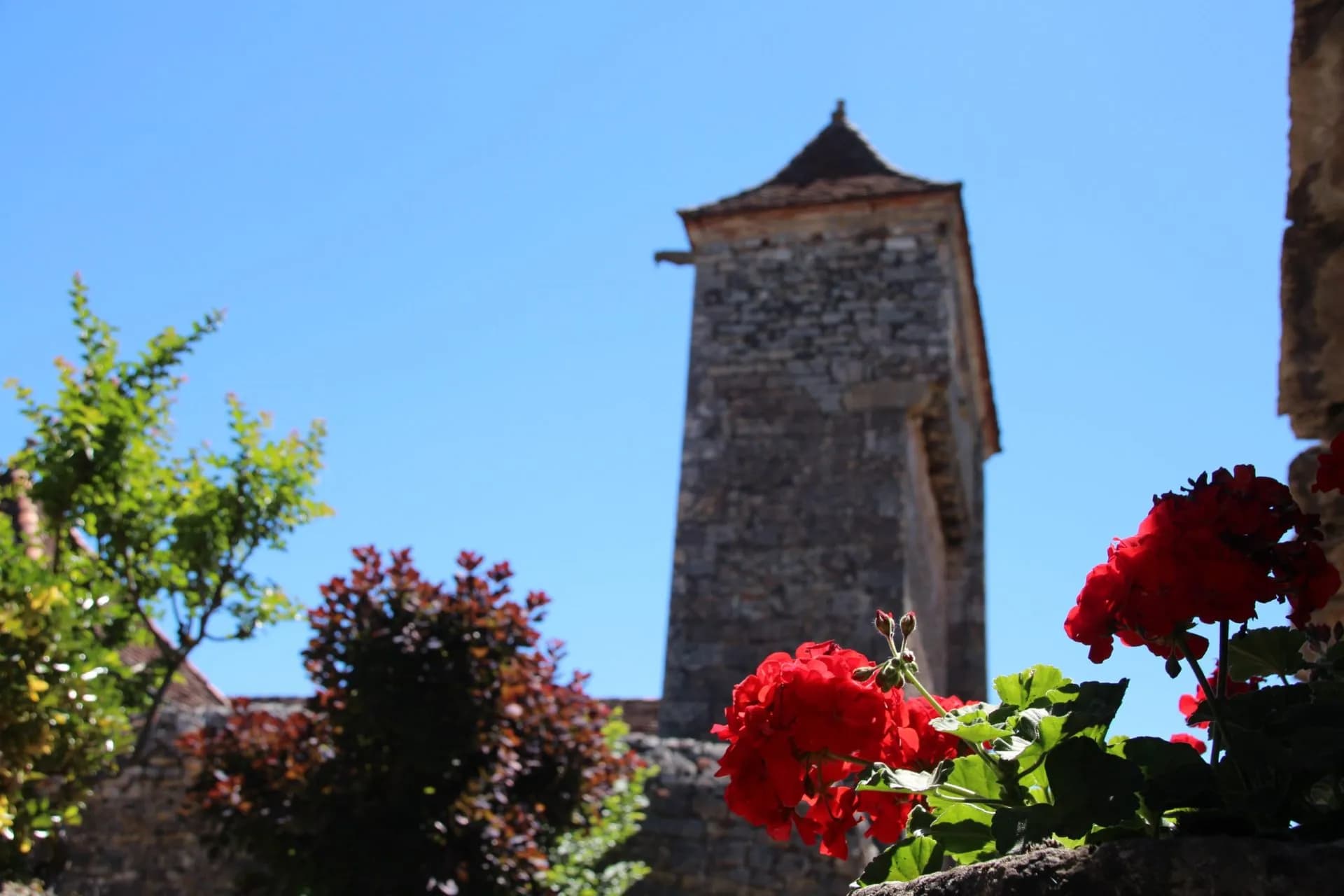 Red geraniums frame a stone tower with a tiled roof under a bright blue sky in Loubressac.