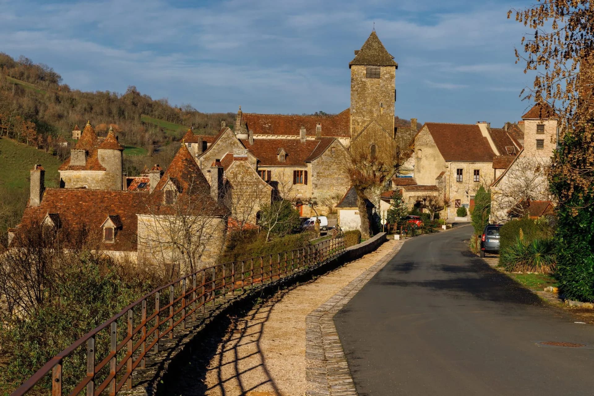 Historic stone village with terracotta roofs and a tall tower, set against a wooded hillside.