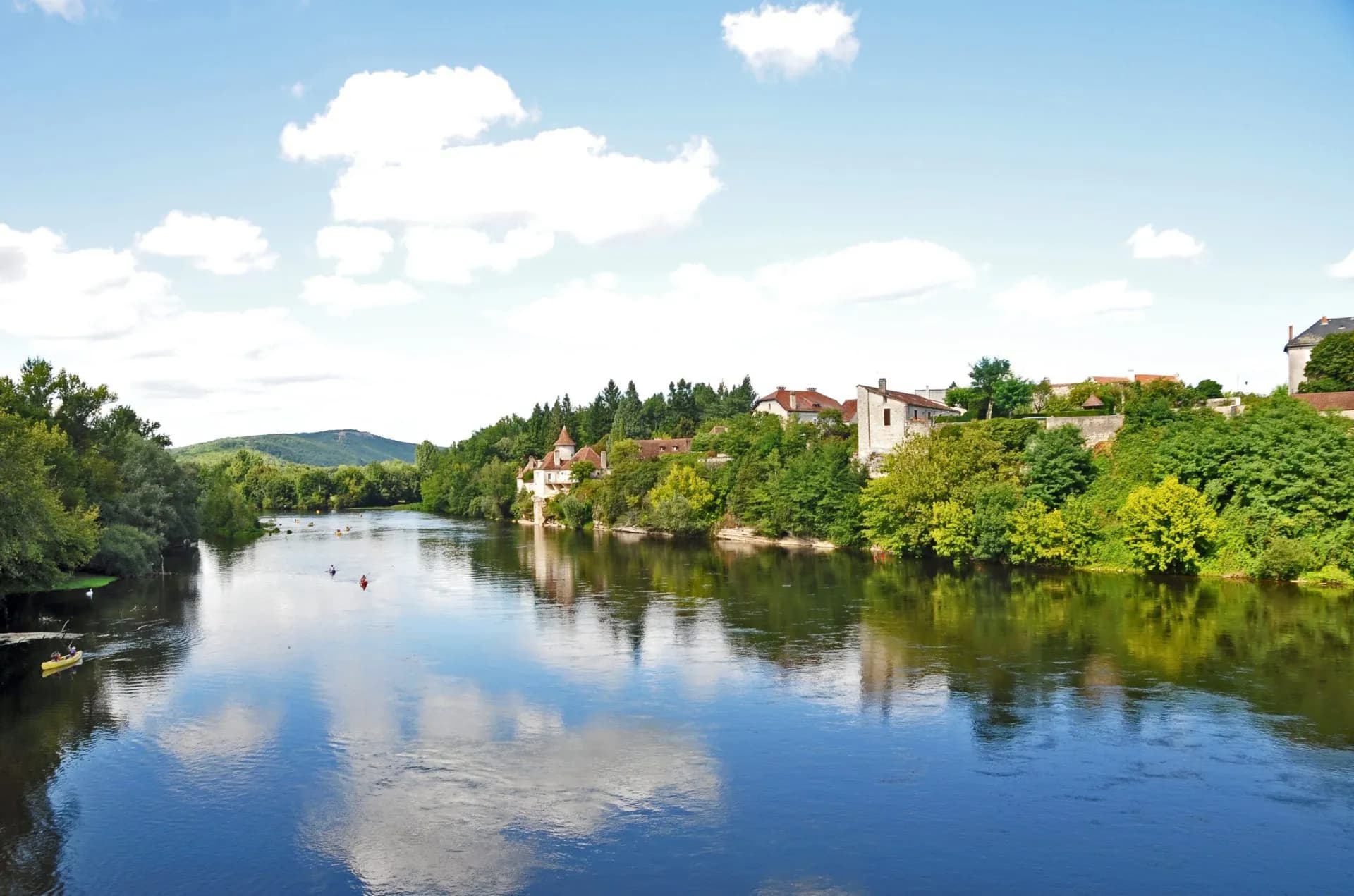 Kayakers paddling on the Dordogne River past riverside buildings under a blue sky.