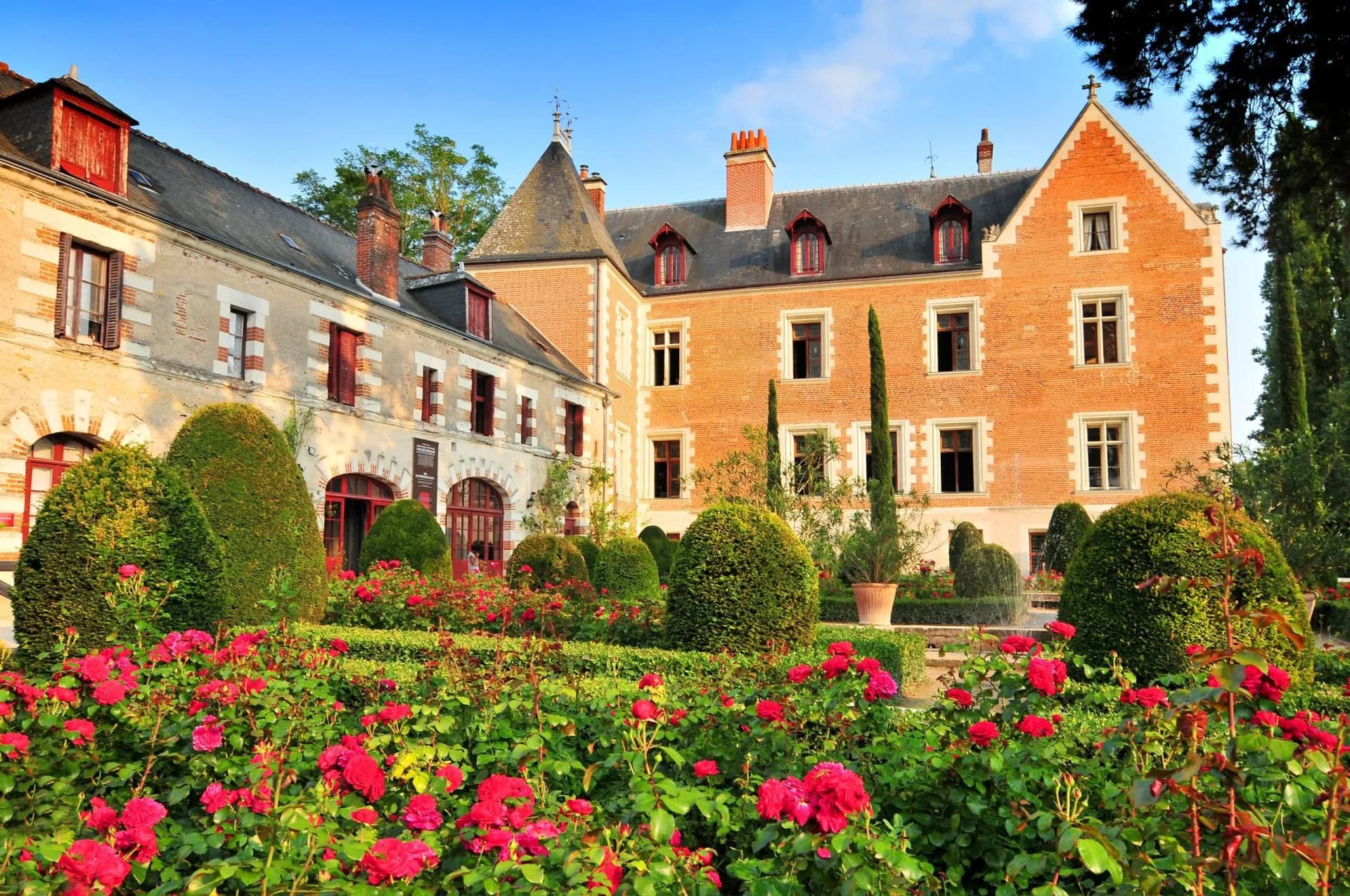 Chateau de Clos Luce with vibrant red roses and manicured topiary gardens under a clear blue sky.