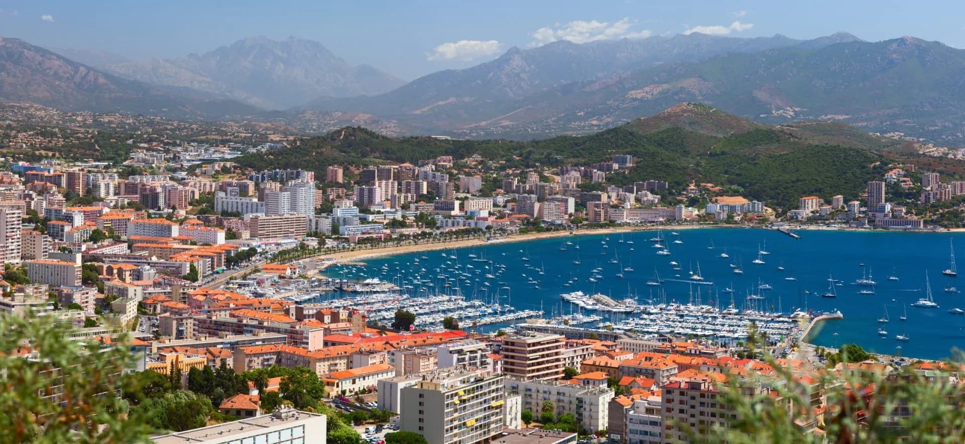 Coastal city of Ajaccio with a harbor full of sailboats against a backdrop of mountains.
