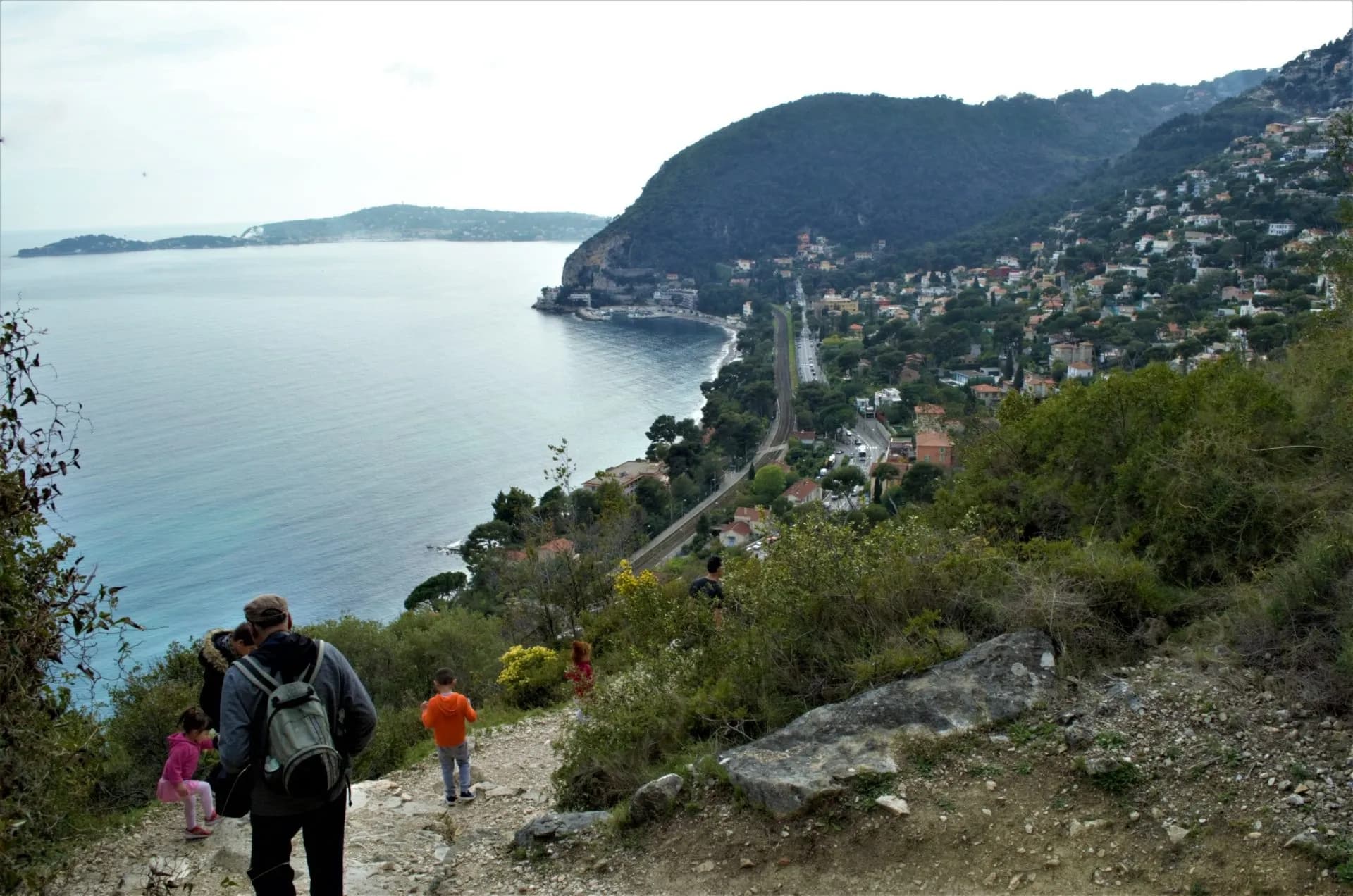 Hikers on dirt trail overlooking Mediterranean coastline and densely built hillside town