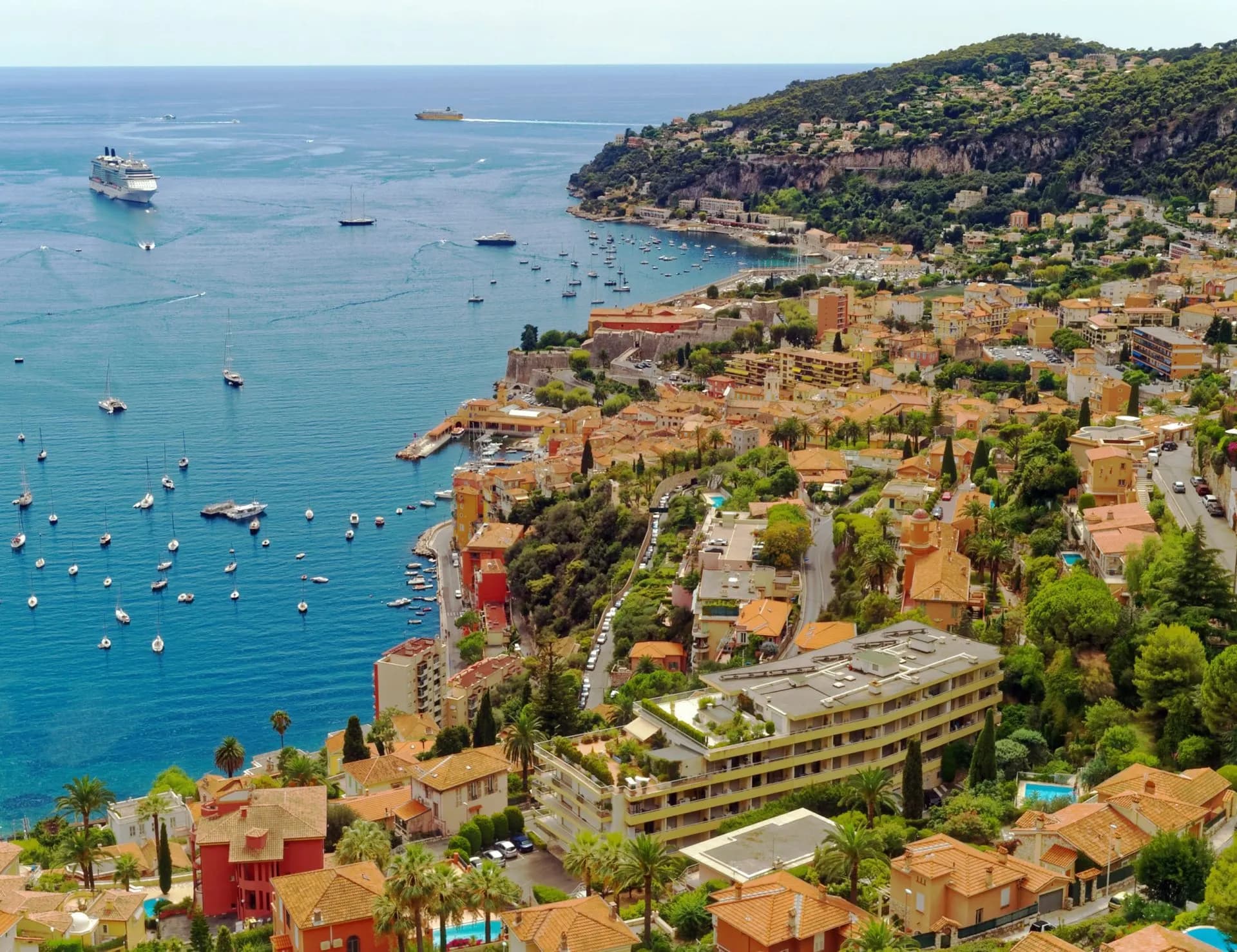 Coastal town with terracotta roofs overlooking the Mediterranean Sea filled with boats and a cruise ship.