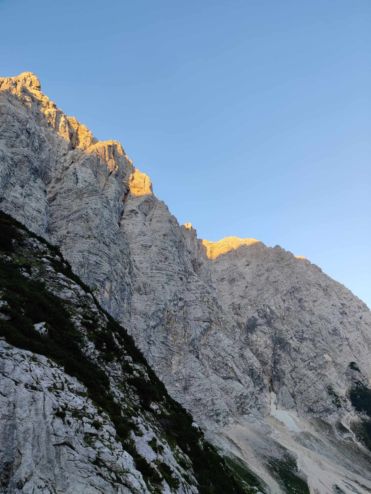 Triglav North Face rock wall illuminated by sunrise against a clear blue sky.