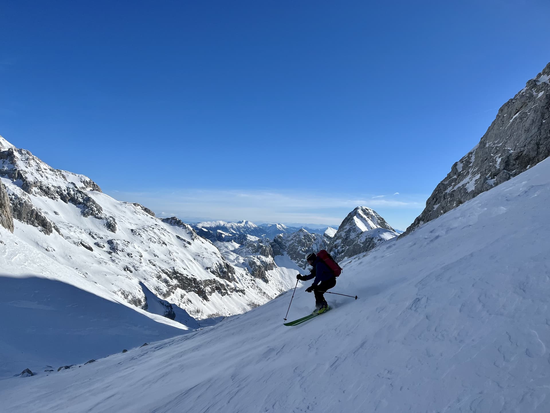 Ski touring down a steep snow slope surrounded by rugged, snow-covered alpine mountains under a clear blue sky.
