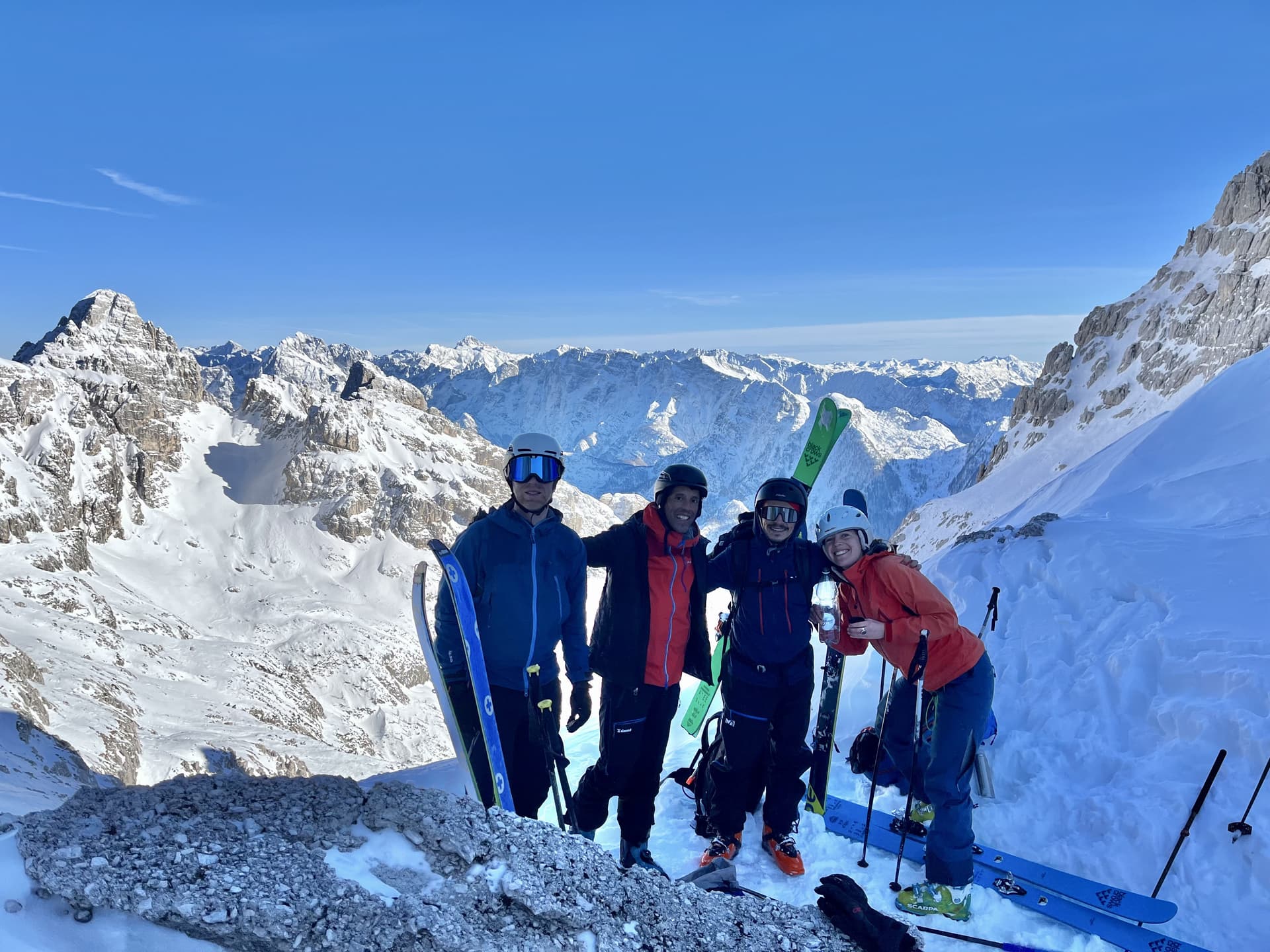 Ski touring group on snowy mountain summit with vast alpine range under clear blue sky.