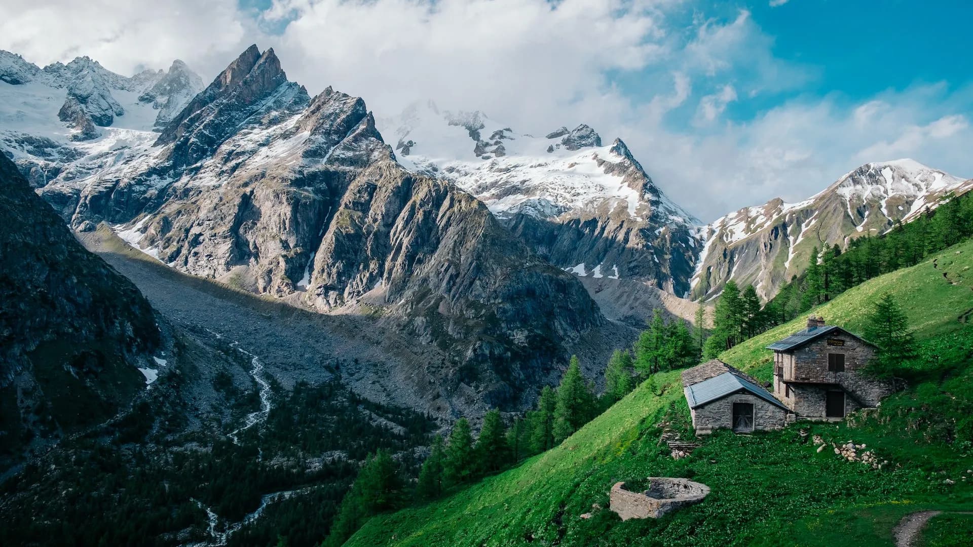 Stone refuge huts on green slope below snow-capped alpine mountains, Refuge Bonatti.