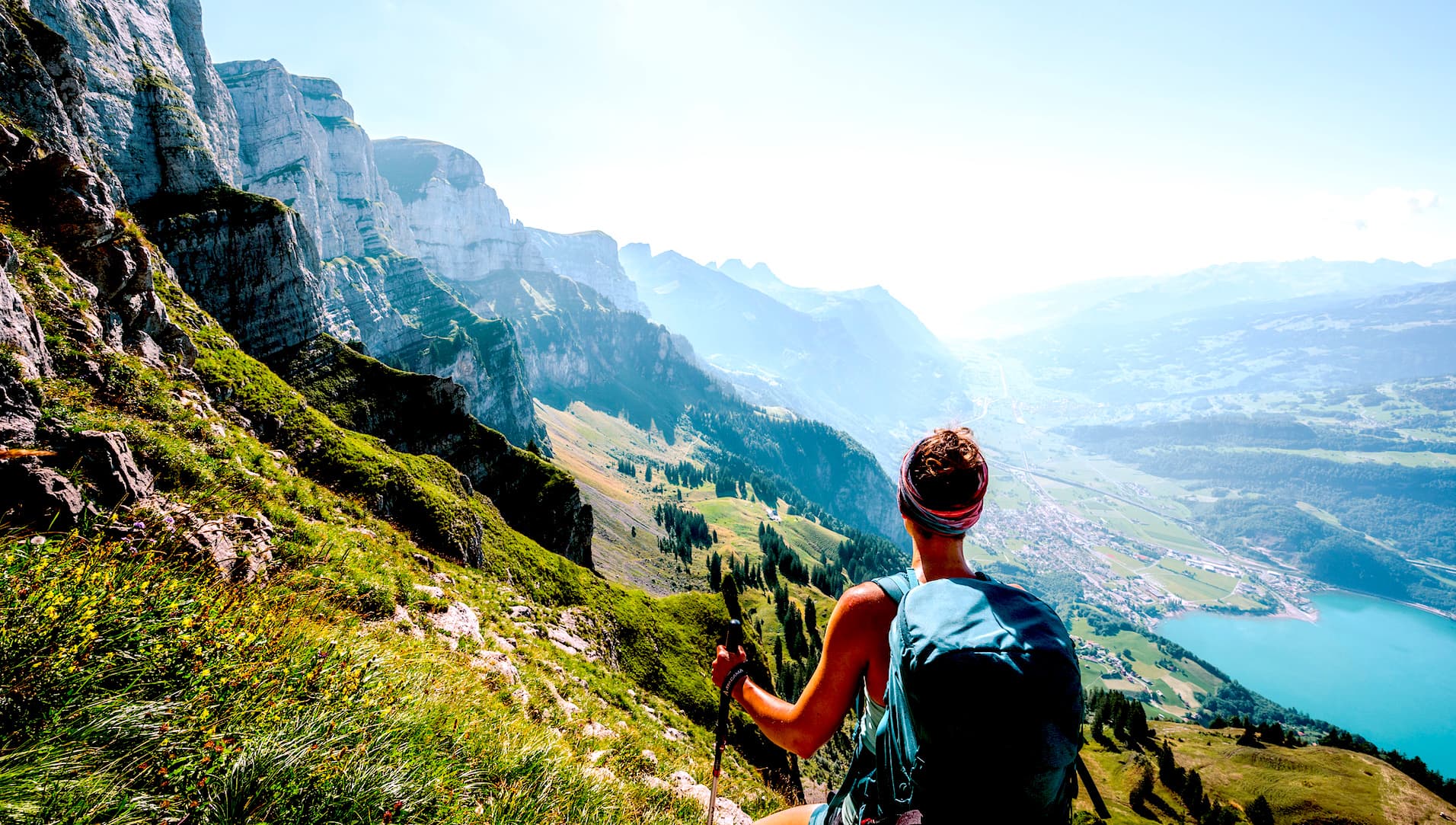 Hiker with backpack and poles overlooking steep green mountains and a valley with a turquoise lake.