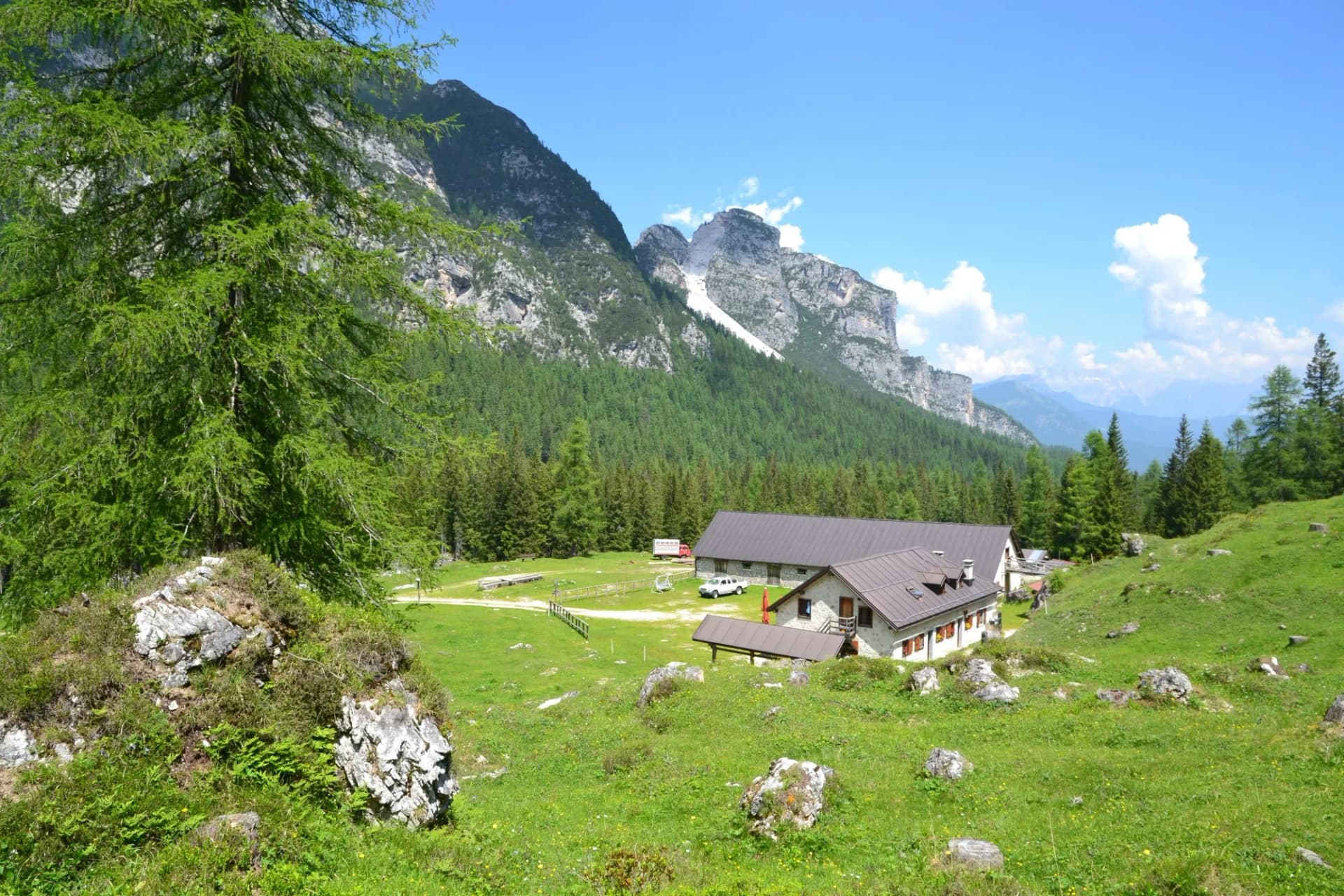 Malga Pramper mountain hut in green alpine meadow with steep Dolomite peaks under blue sky.
