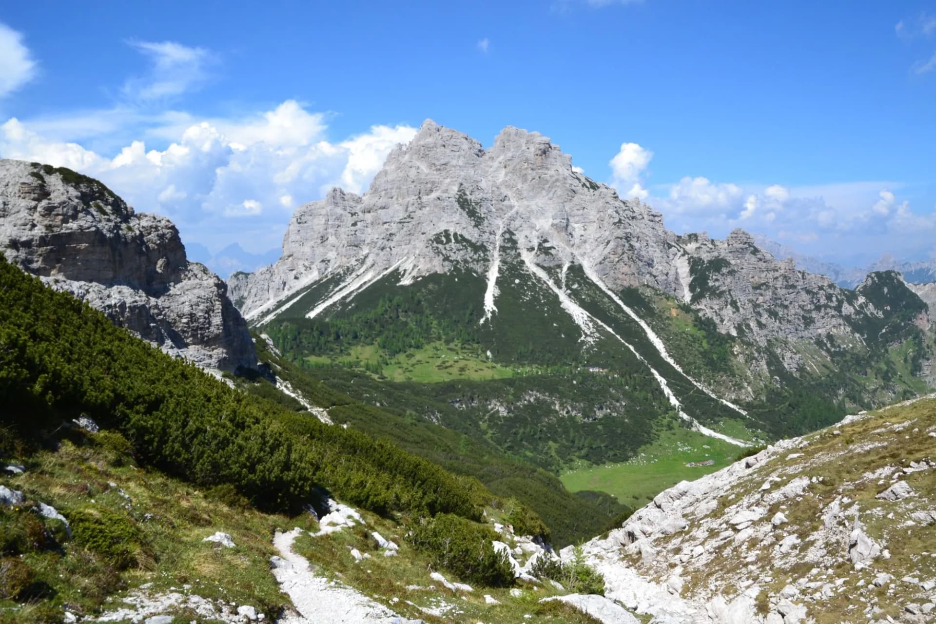Rocky mountain peaks above green slopes and a hiking trail under a blue sky with clouds.