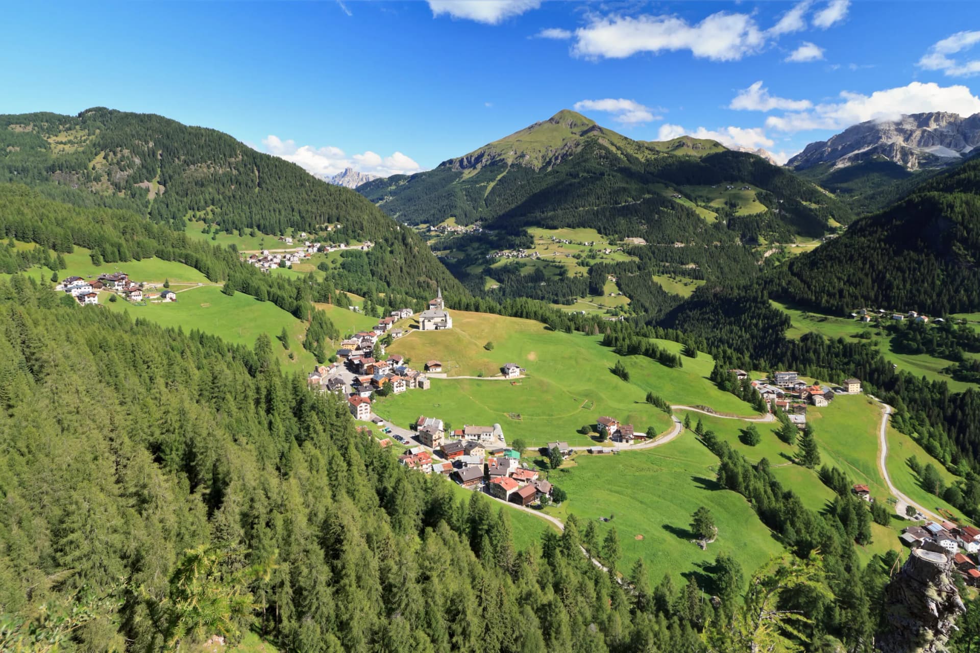 Alpine village nestled in green valley with steep forested slopes and rocky peaks under blue sky.
