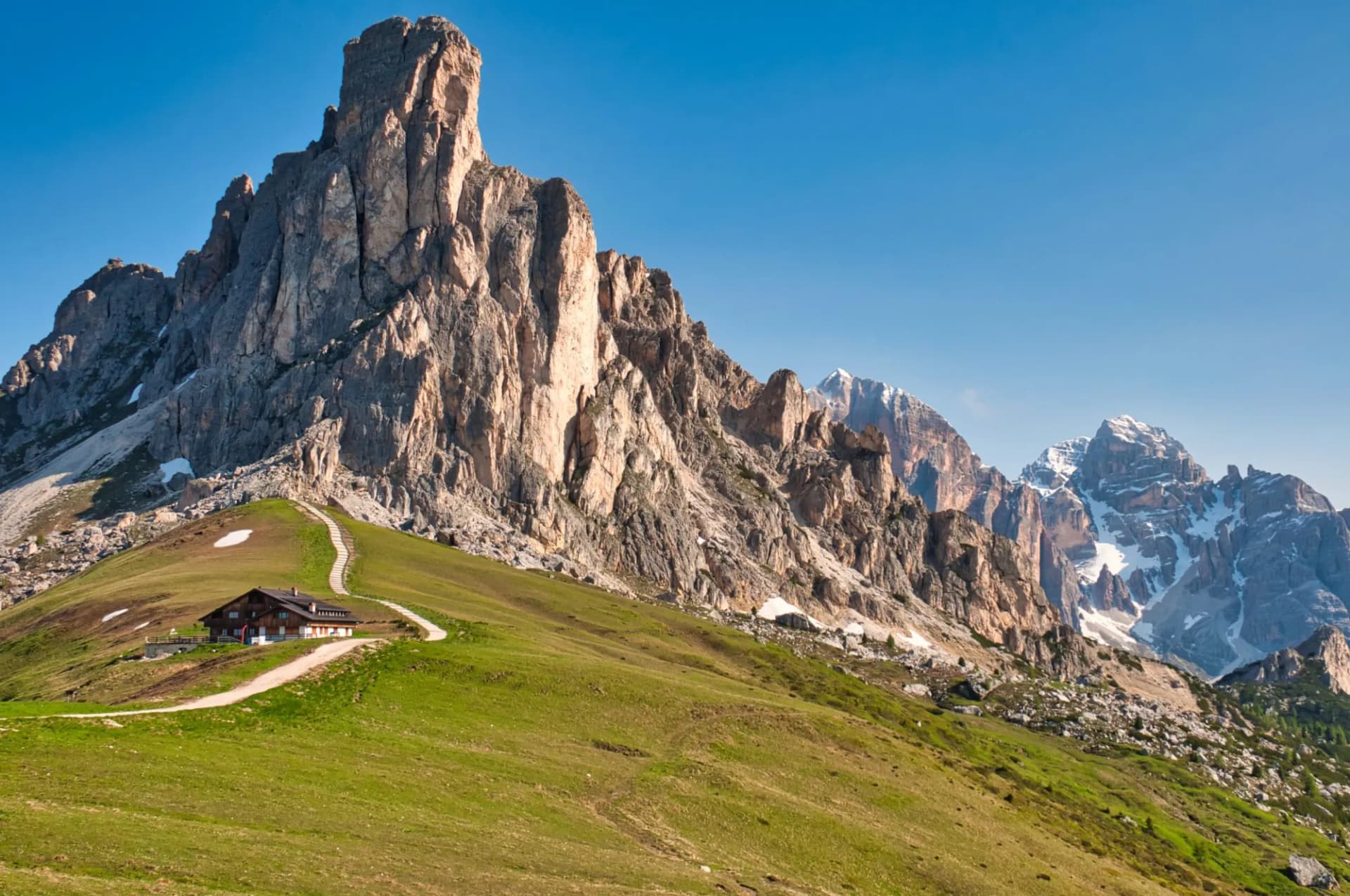 Mountain hut on grassy slope below Passo Giau peaks with snow-capped mountains in background