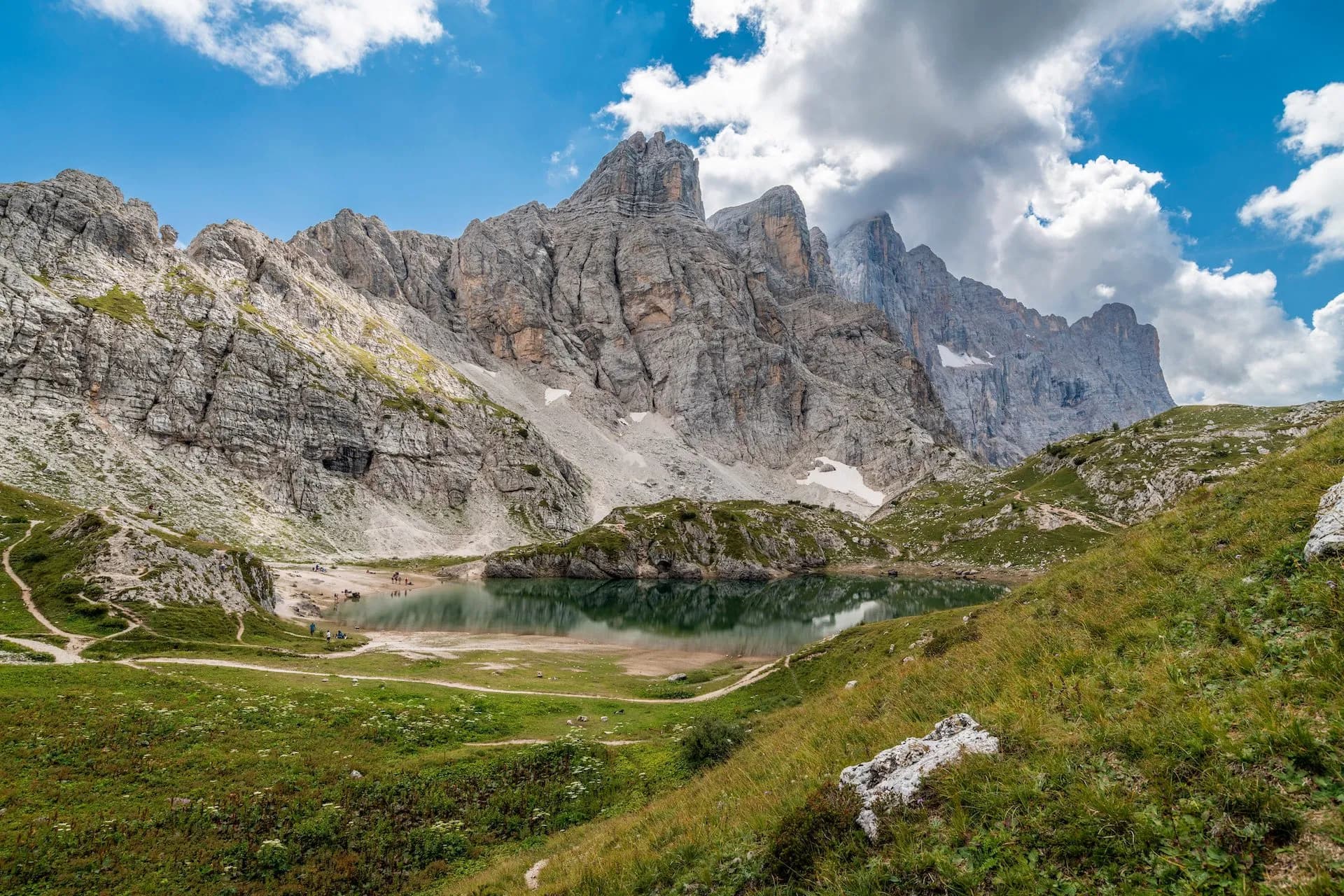 Alpine lake reflecting jagged grey mountains under a blue and cloudy sky, with green meadows.