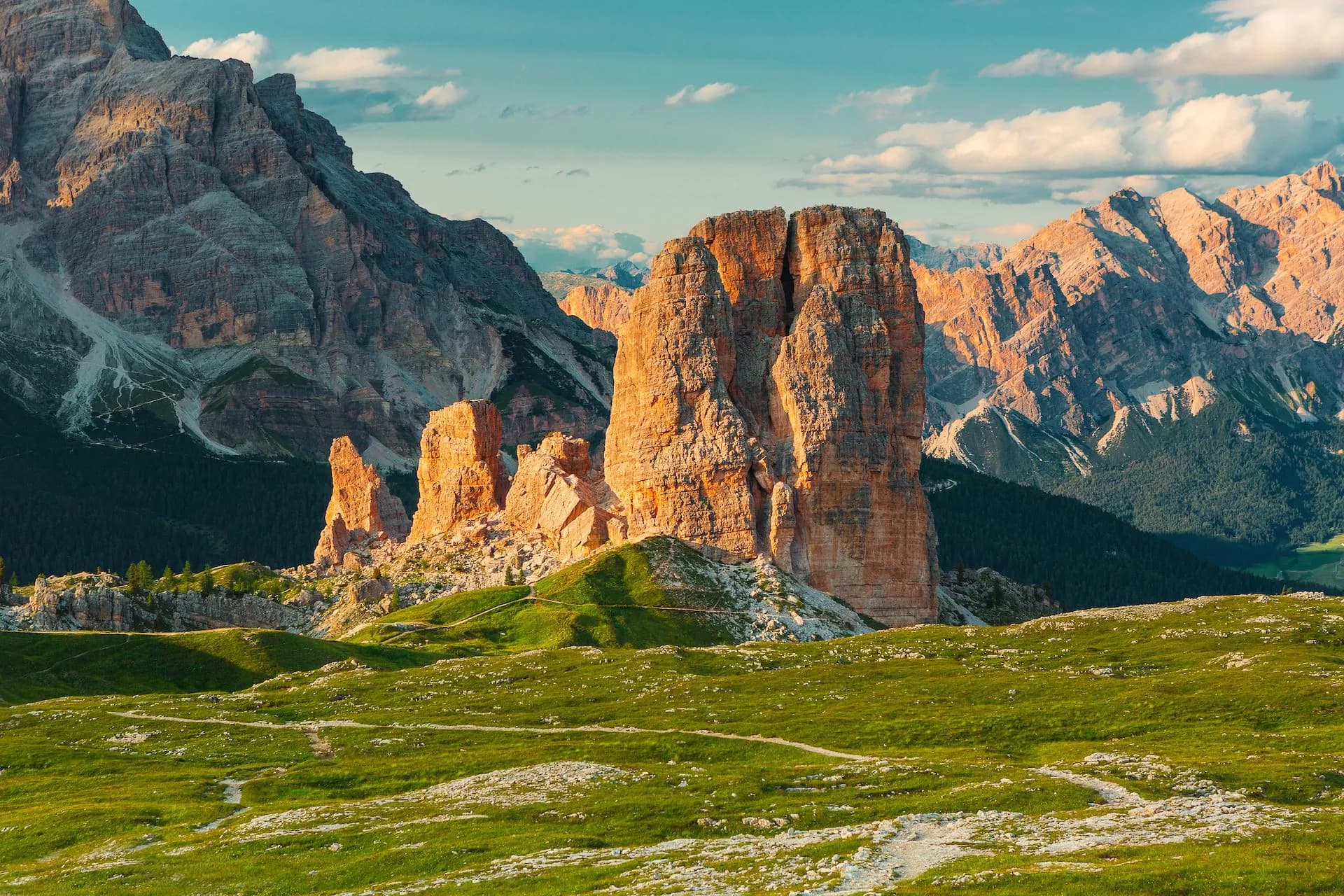 Cinque Torri rock formations illuminated by sun above green alpine meadow with hiking trails.