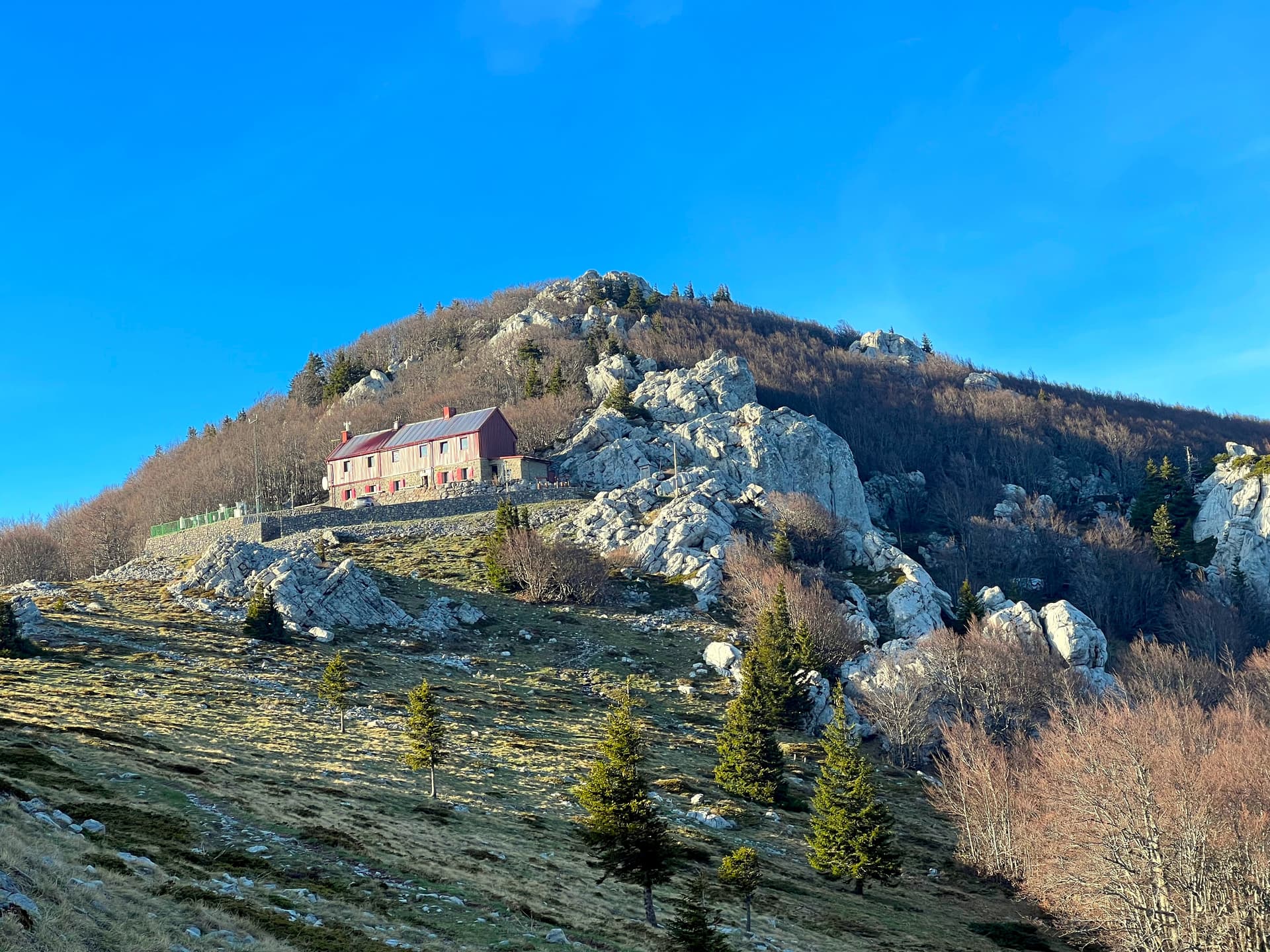 Mountain hut on rocky slope with sparse trees under a clear blue sky, Zavizan Hut.