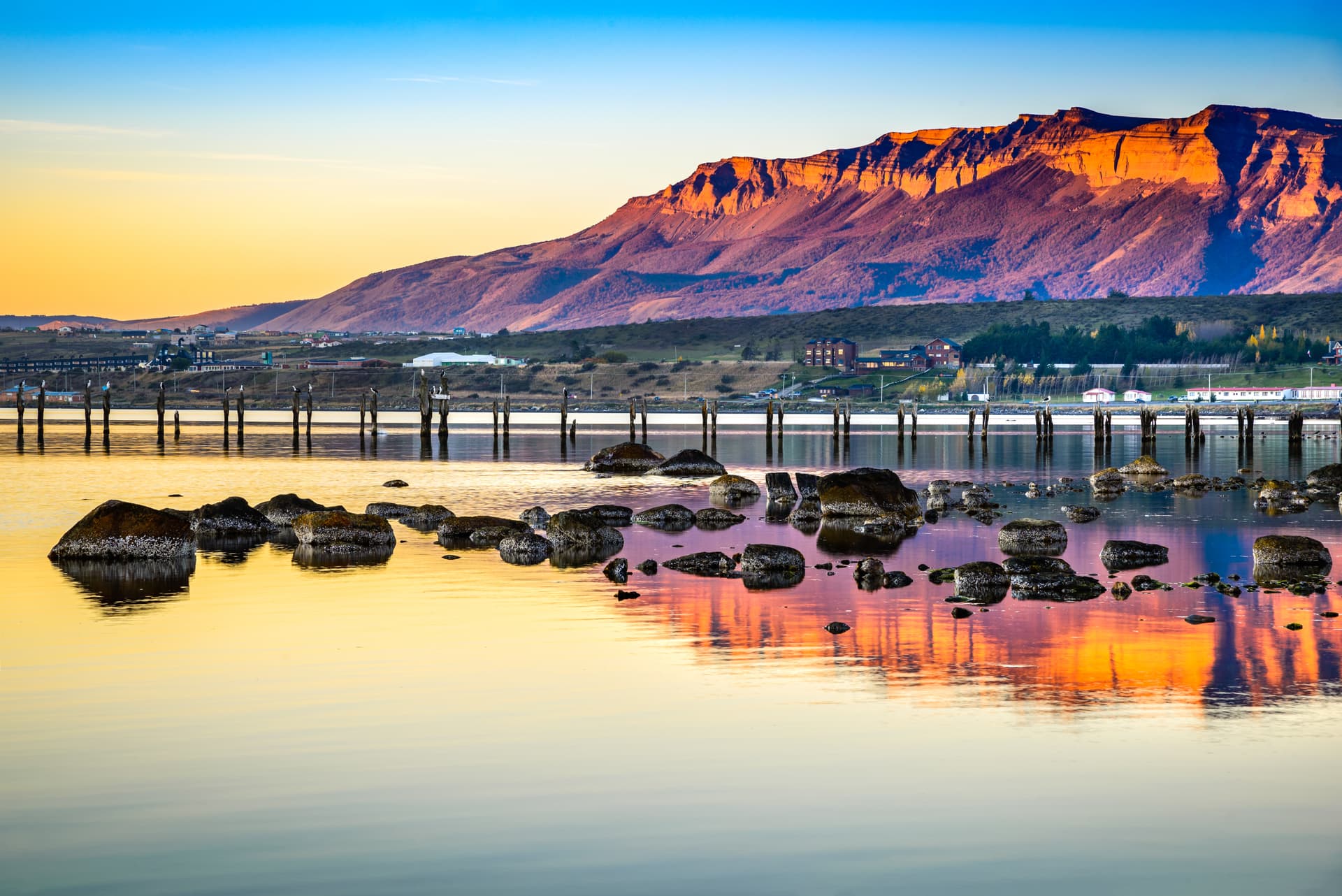 Mountains reflecting sunset over water with rocks and pier pilings in Puerto Natales, Chile.