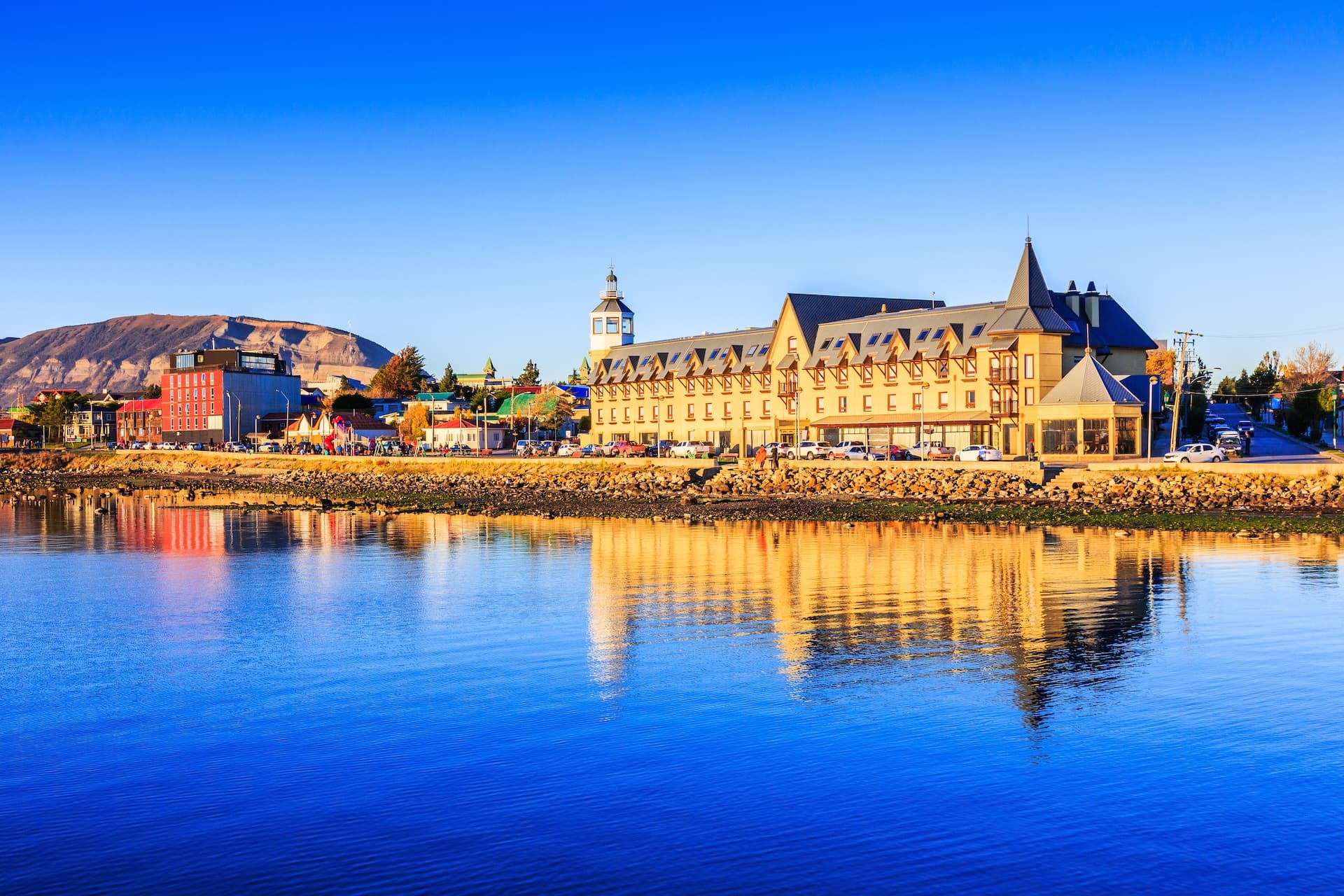 Puerto Natales waterfront buildings reflecting in deep blue water with mountains in background.