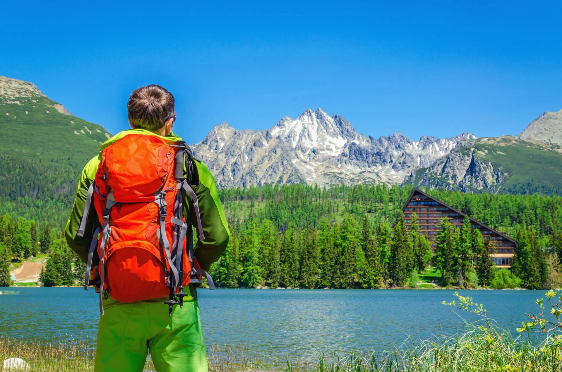 Hiker with orange backpack overlooking Štrbské Pleso lake and snowy Tatra Mountains.