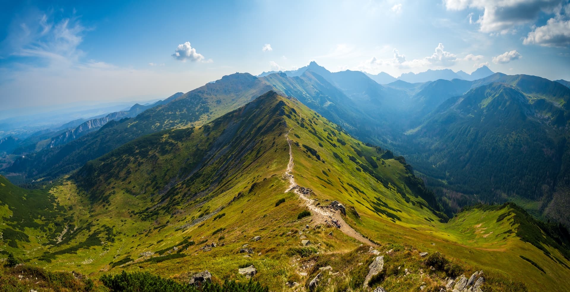 Hiking trail along a grassy mountain ridge with distant peaks under a blue sky