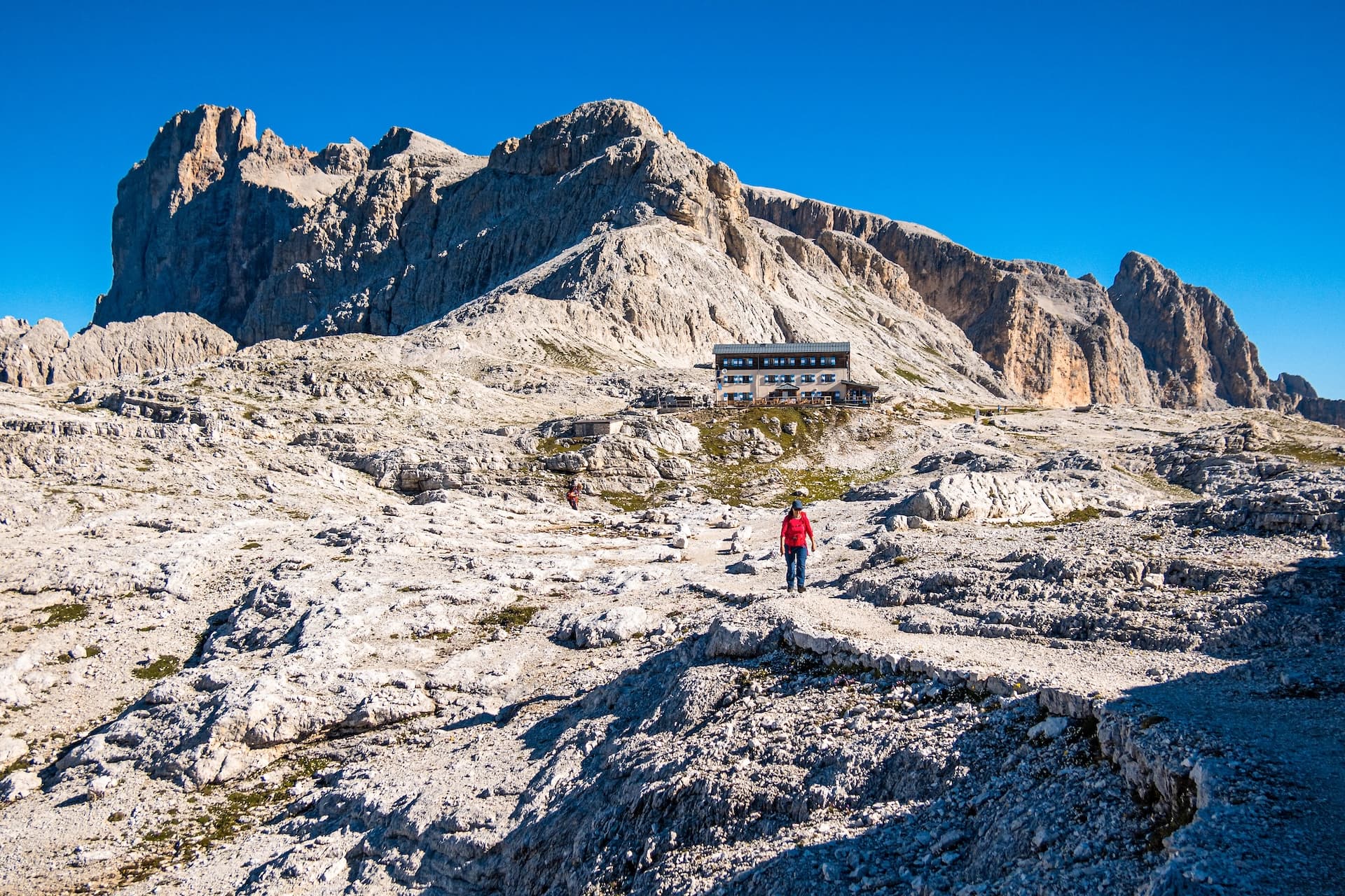 Hiker on rocky terrain approaching Rifugio Rosetta below massive Dolomite mountain peaks.