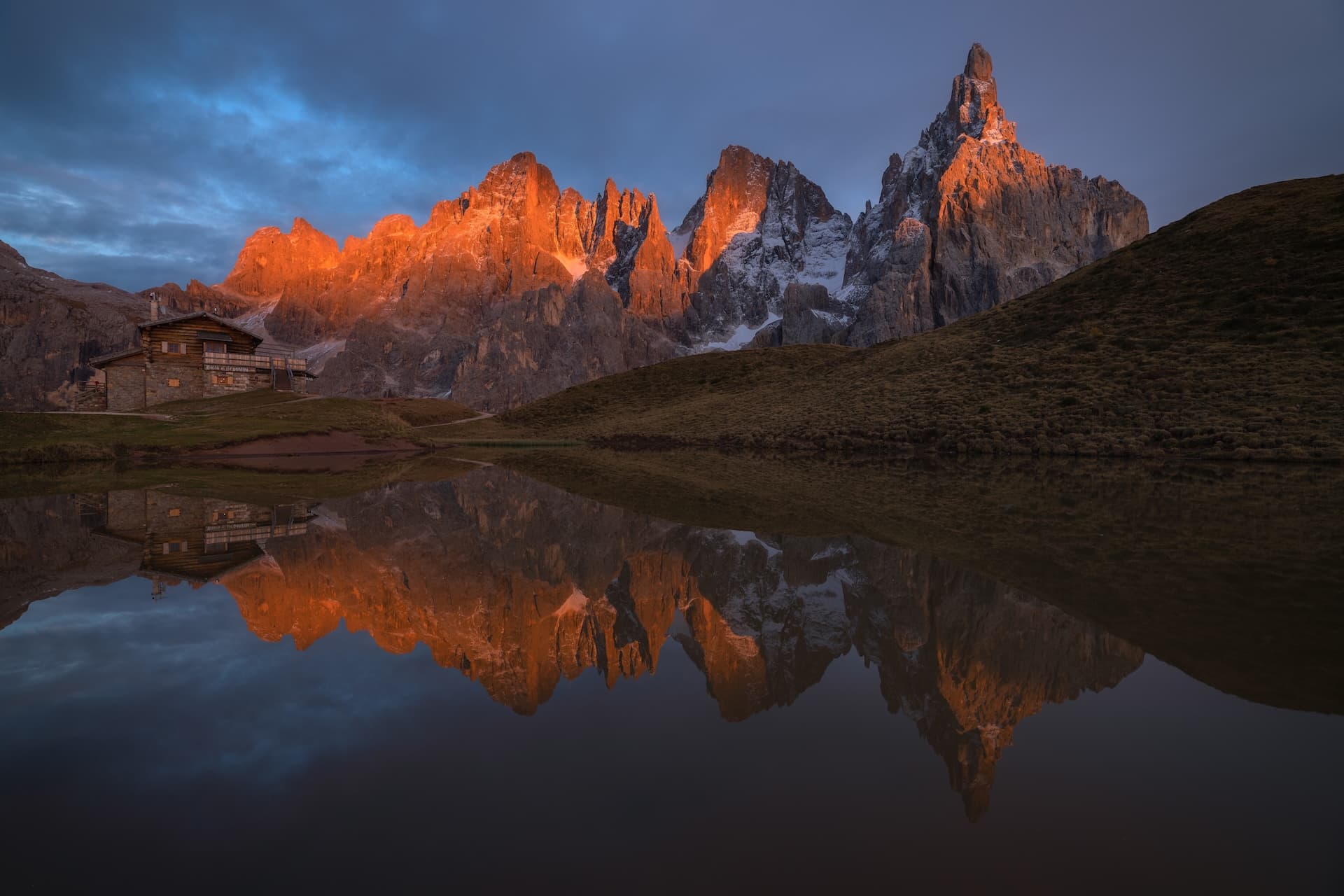 Alpenglow illuminates rugged mountain peaks reflected in a calm alpine lake near a wooden cabin.