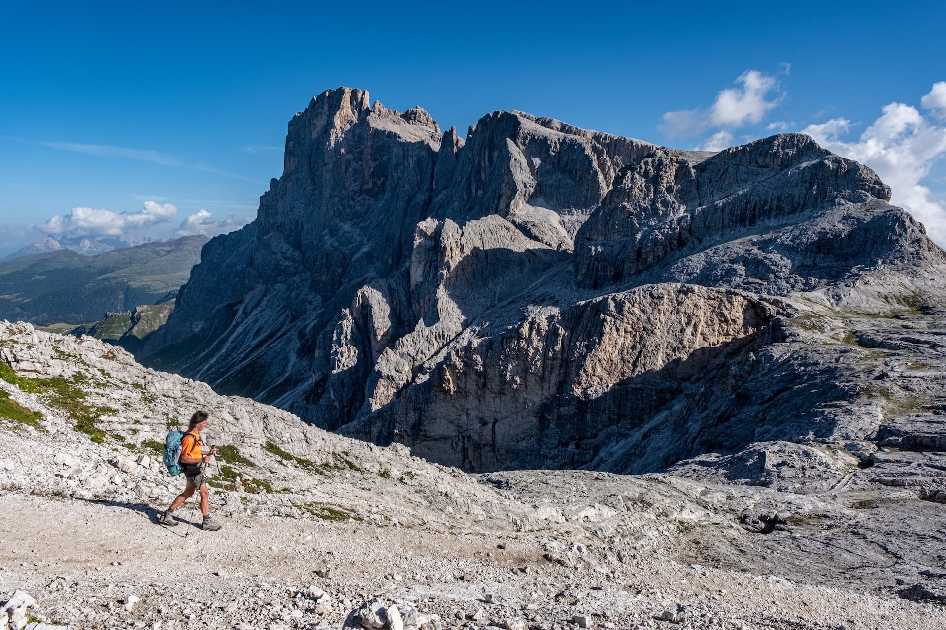 Hiker with backpack walking on rocky trail below massive gray mountain peaks under blue sky.