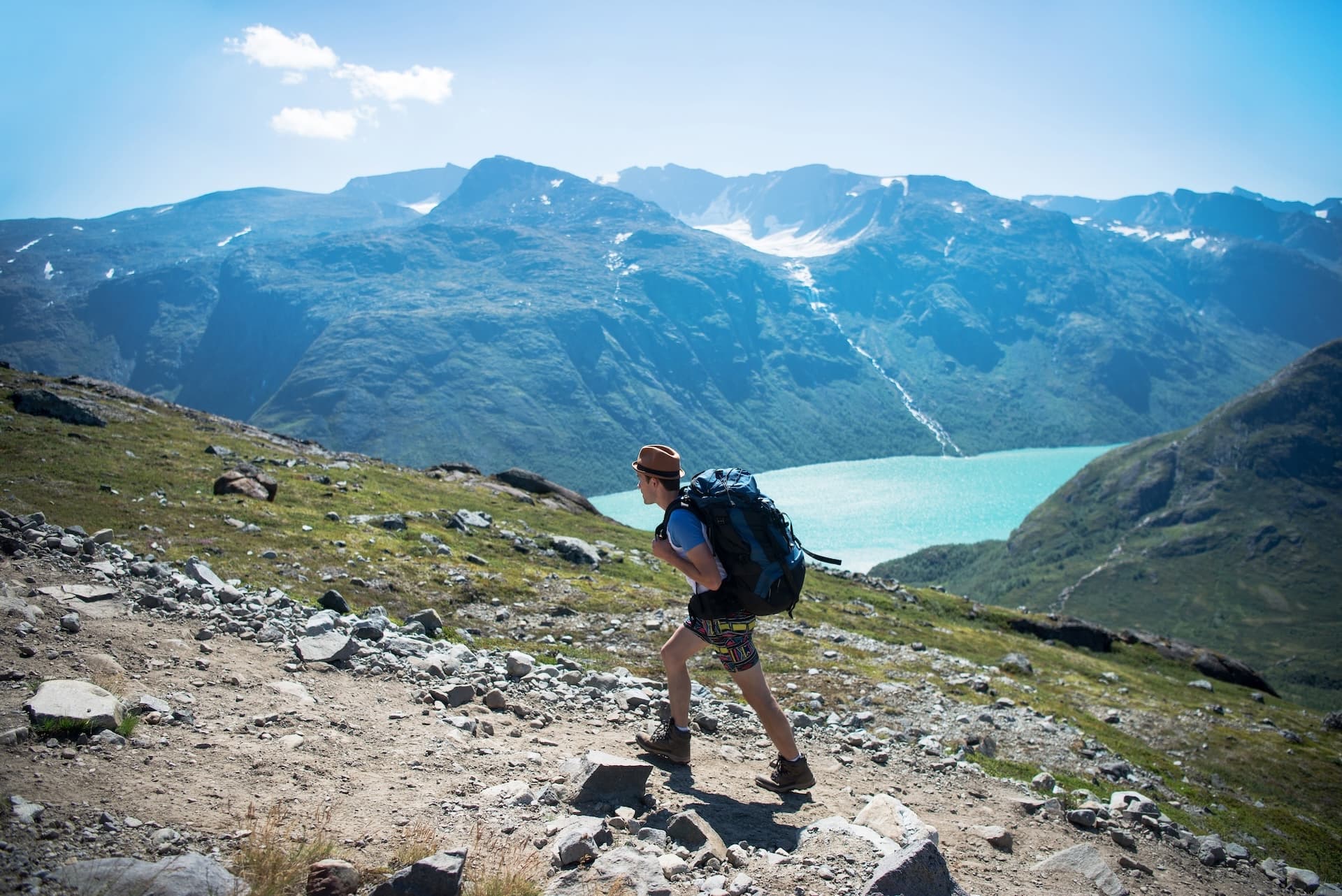 Hiker with backpack ascending rocky trail above Gjende See with turquoise water and mountains.