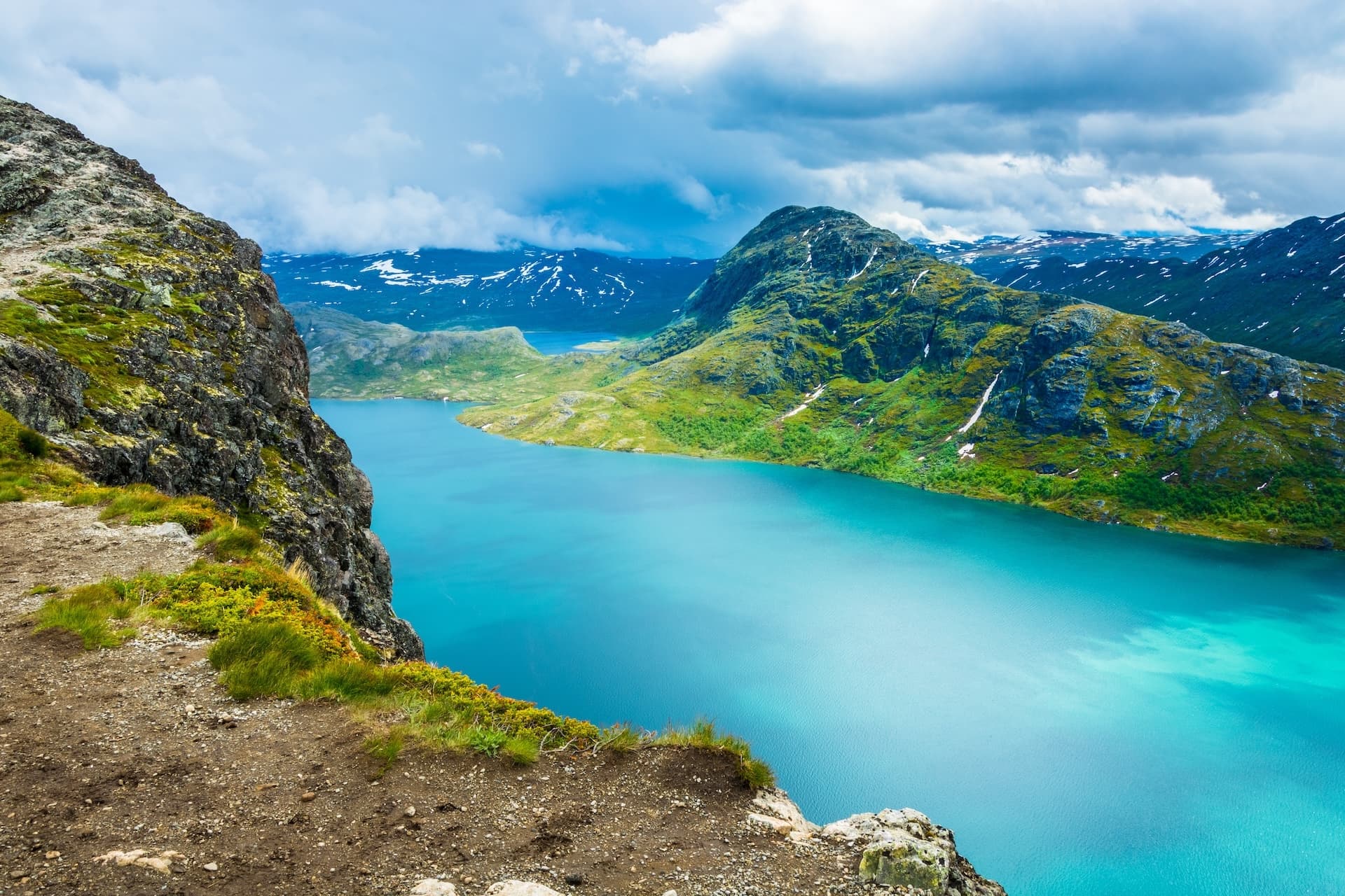 Hiking view from Besseggen Ridge overlooking bright turquoise lake and green mountains with snow.