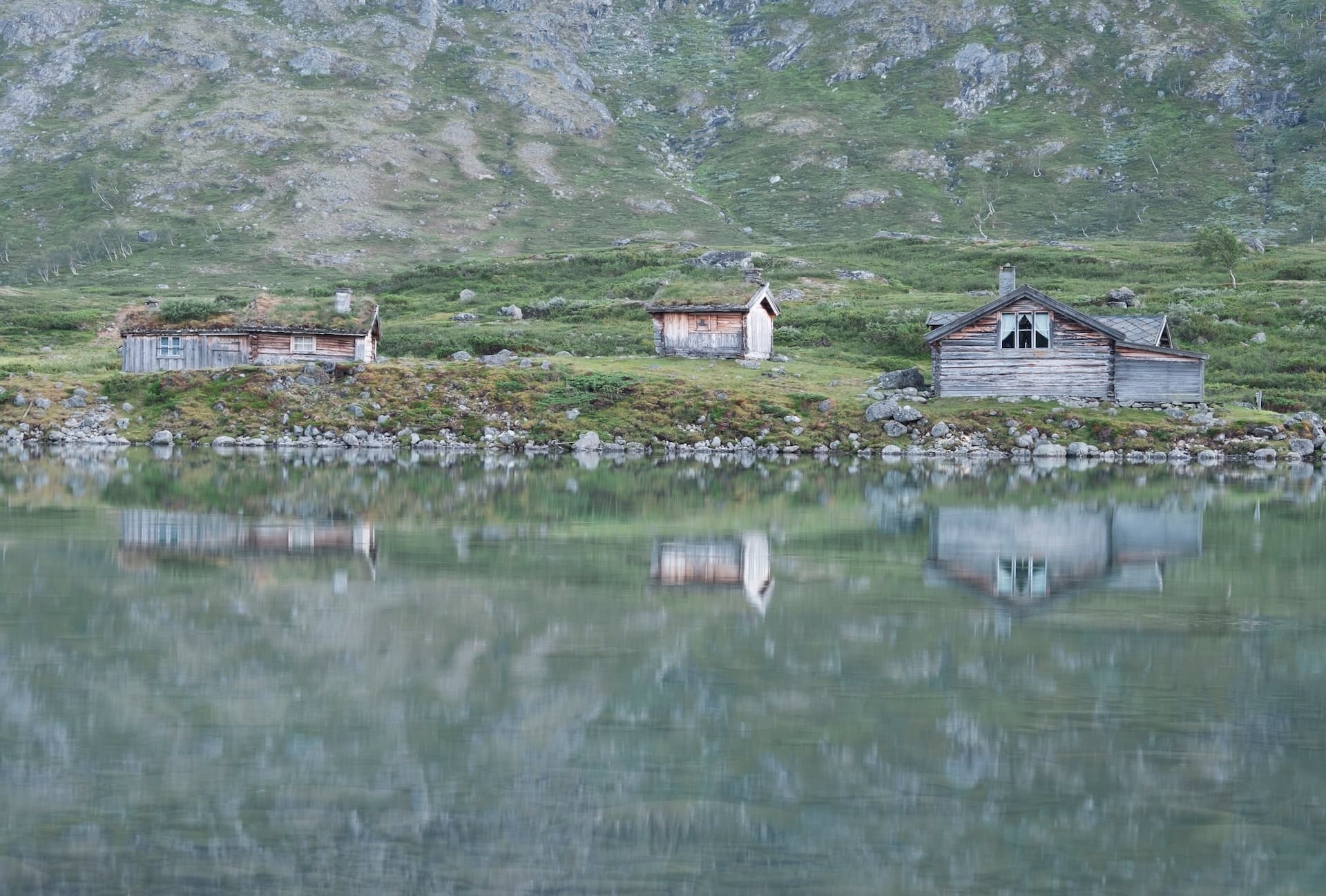 Wooden huts with turf roofs reflected in Gjende lake against a grassy mountain slope.