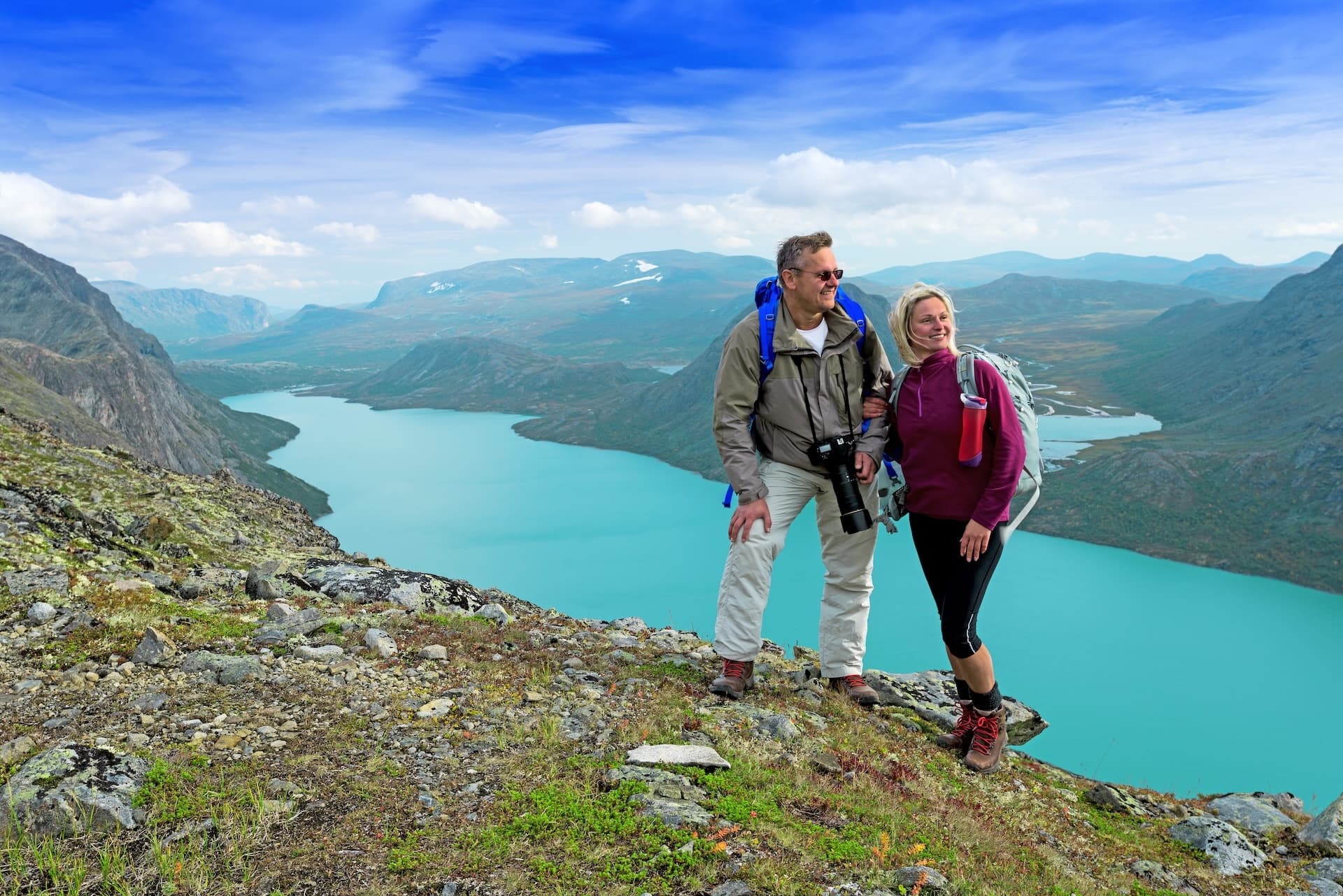 Hikers with backpacks stand on rocky terrain overlooking a turquoise lake and mountains, Besseggen Ridge.