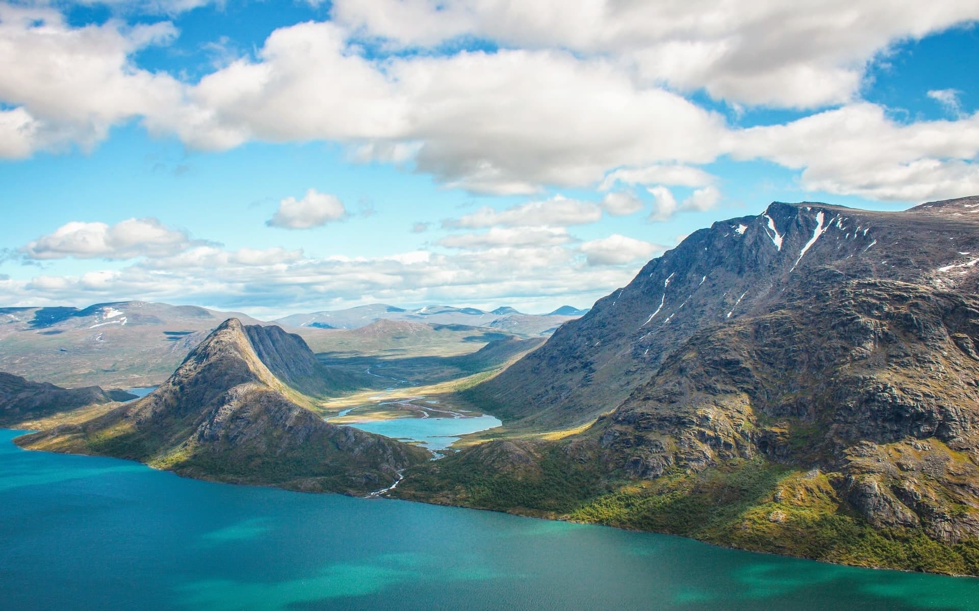 Vast mountain valley with turquoise lakes and patches of snow under a cloudy blue sky in Norway.