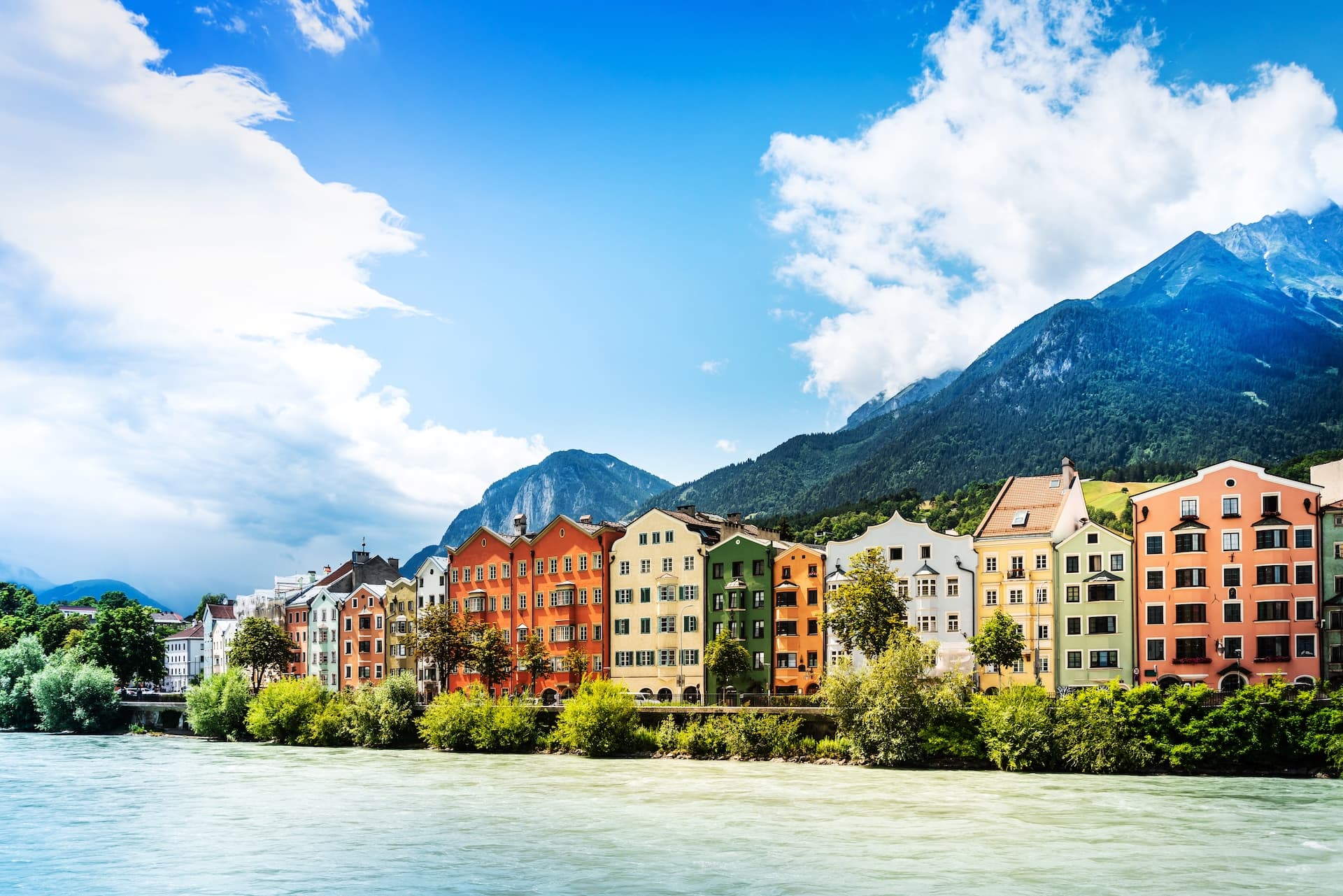Colorful houses along the river in Innsbruck with dramatic green mountains under a blue sky.
