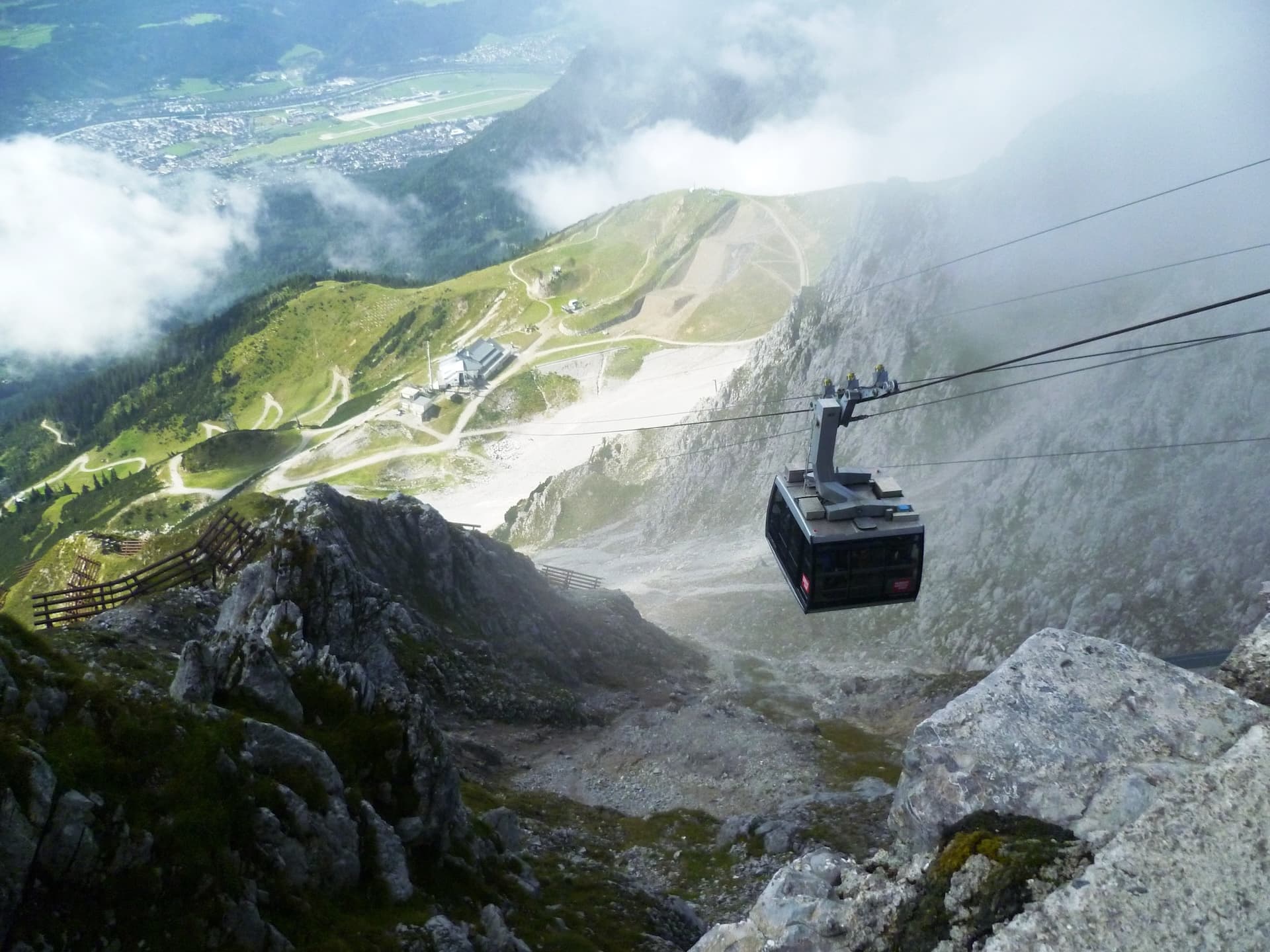 Cable car ascending steep rocky mountains above green slopes and a valley town with an airport.