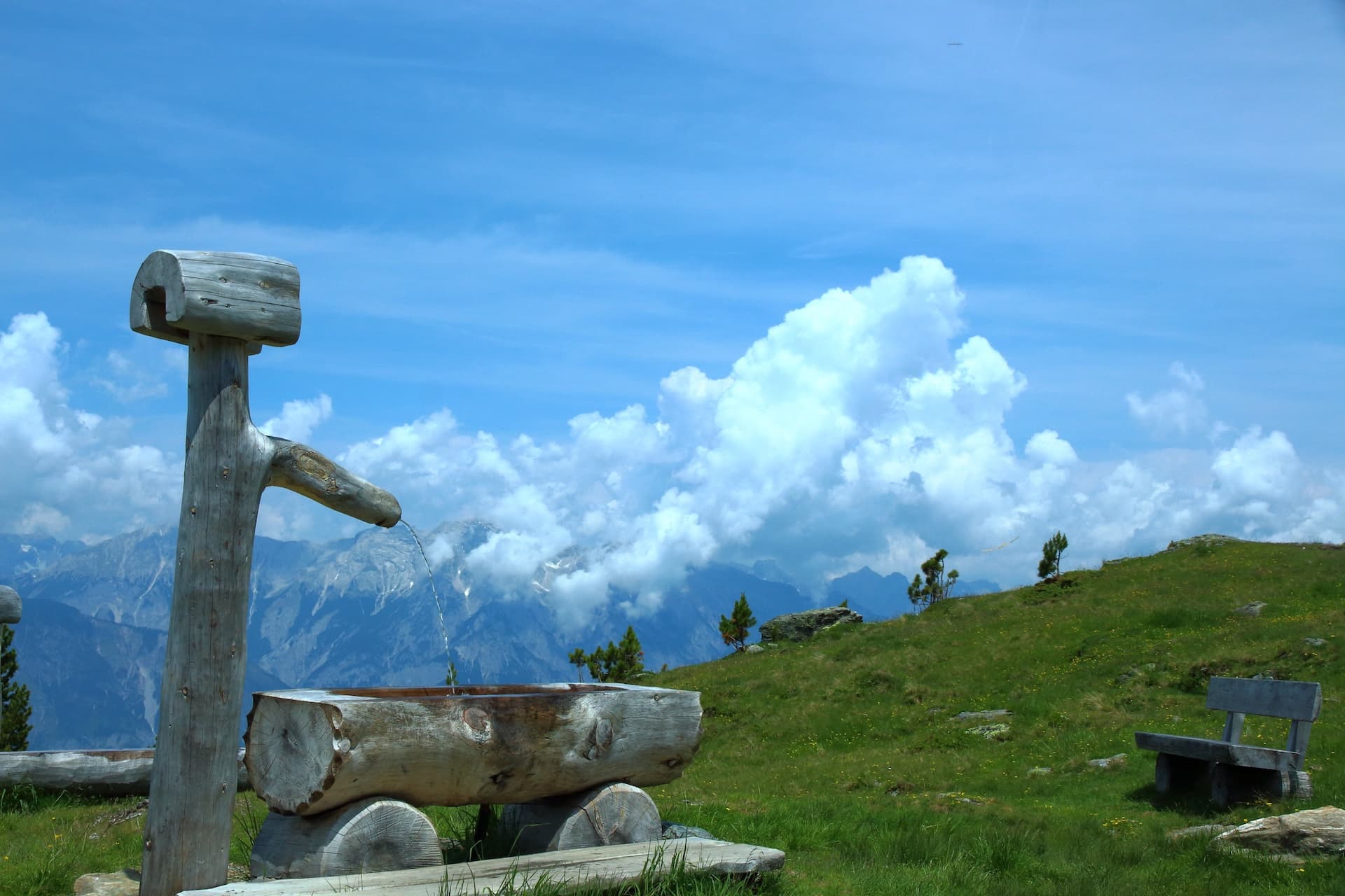 Wooden water trough fountain on grassy mountain slope overlooking blue peaks under cloudy sky