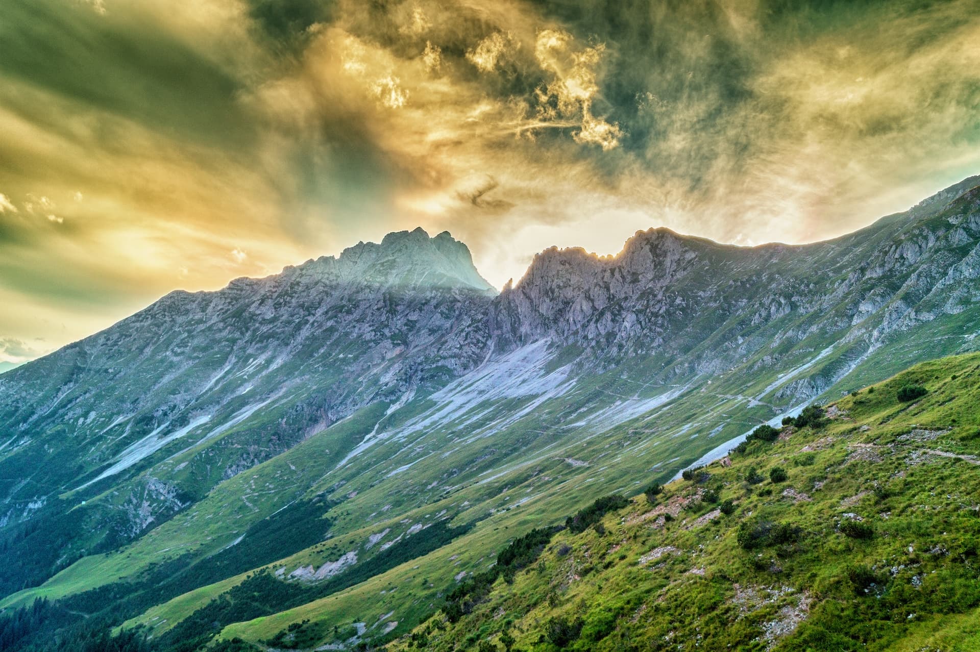 Rugged mountainsides with green slopes under dramatic, golden-hued clouds at sunset