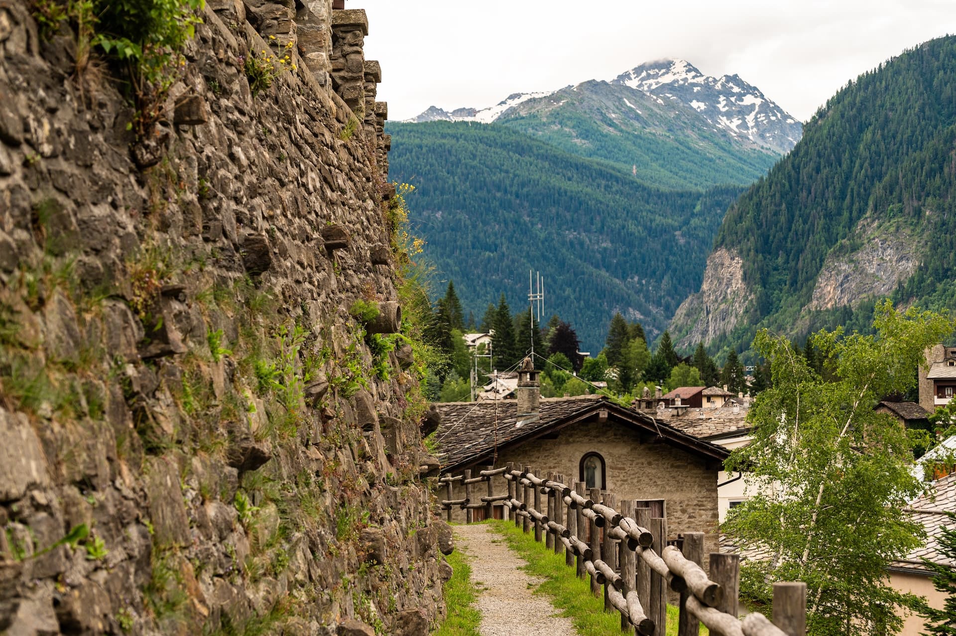 Gravel path beside stone wall in Courmayeur valley with snow-capped mountains.