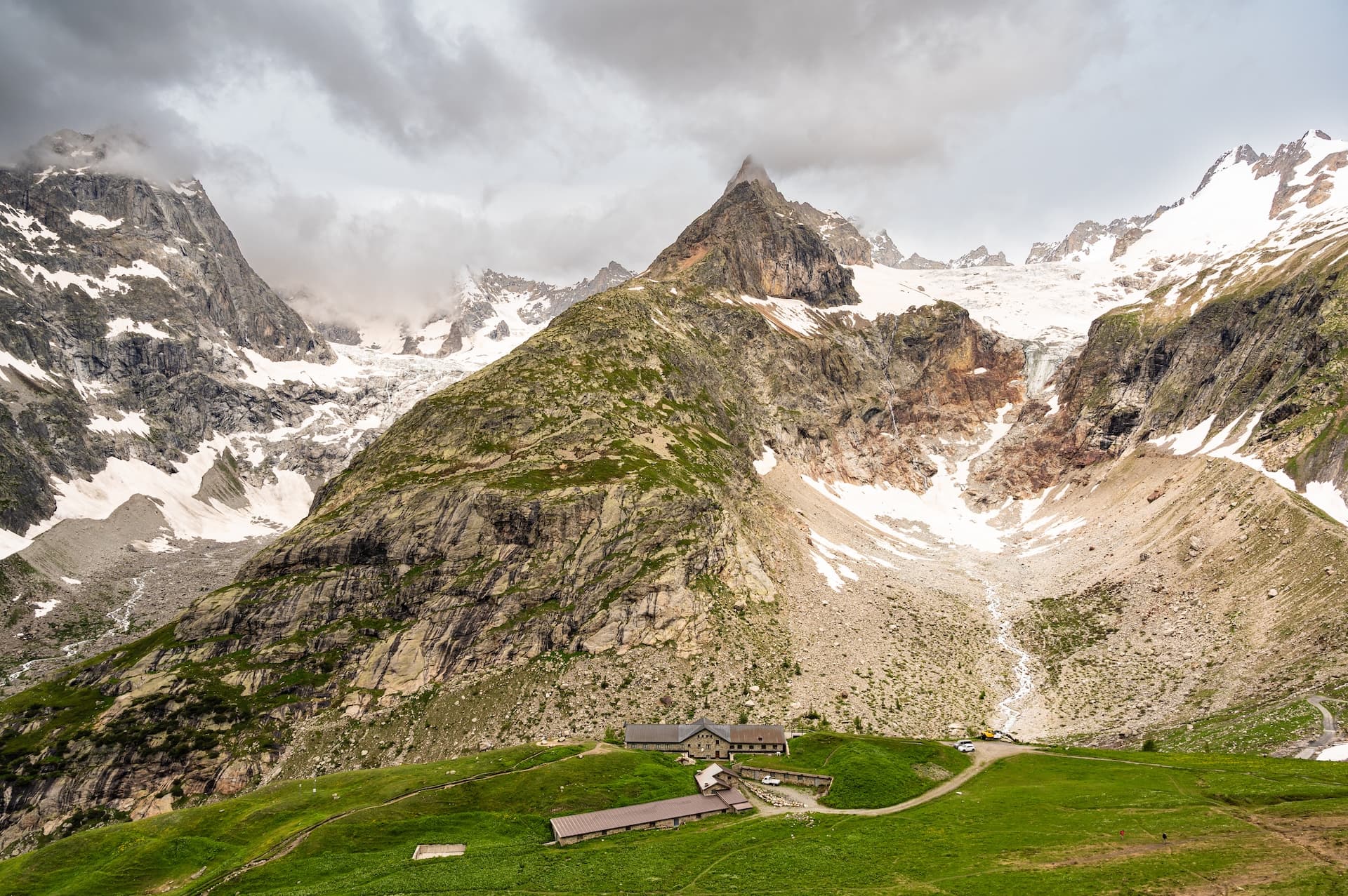 Mountain refuge cottage nestled in green alpine valley below snow-capped peaks and glacier.