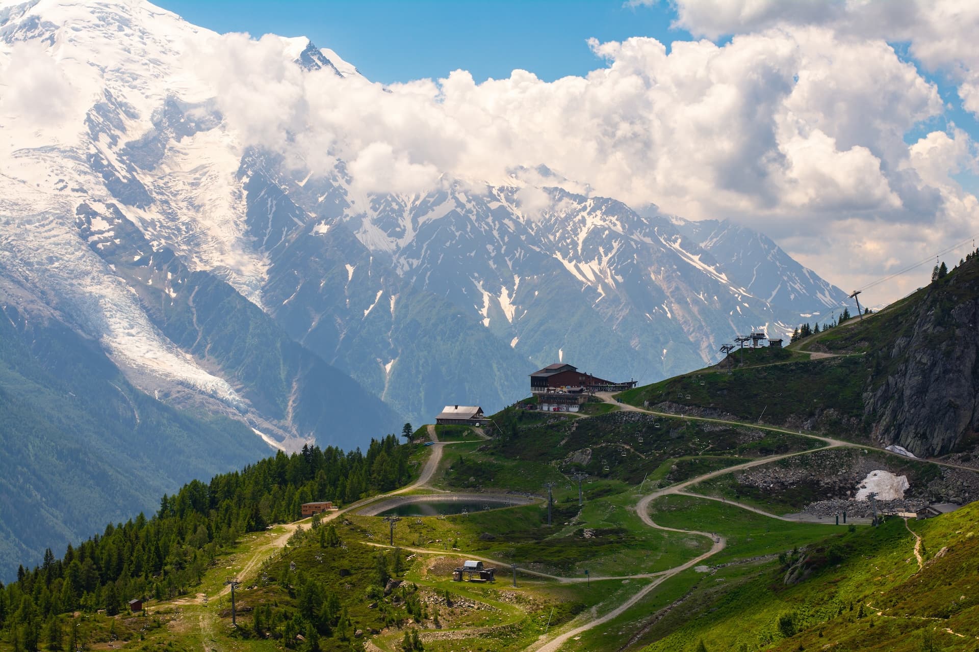 Mountain hut on green slope with winding paths below snow-capped peaks and clouds
