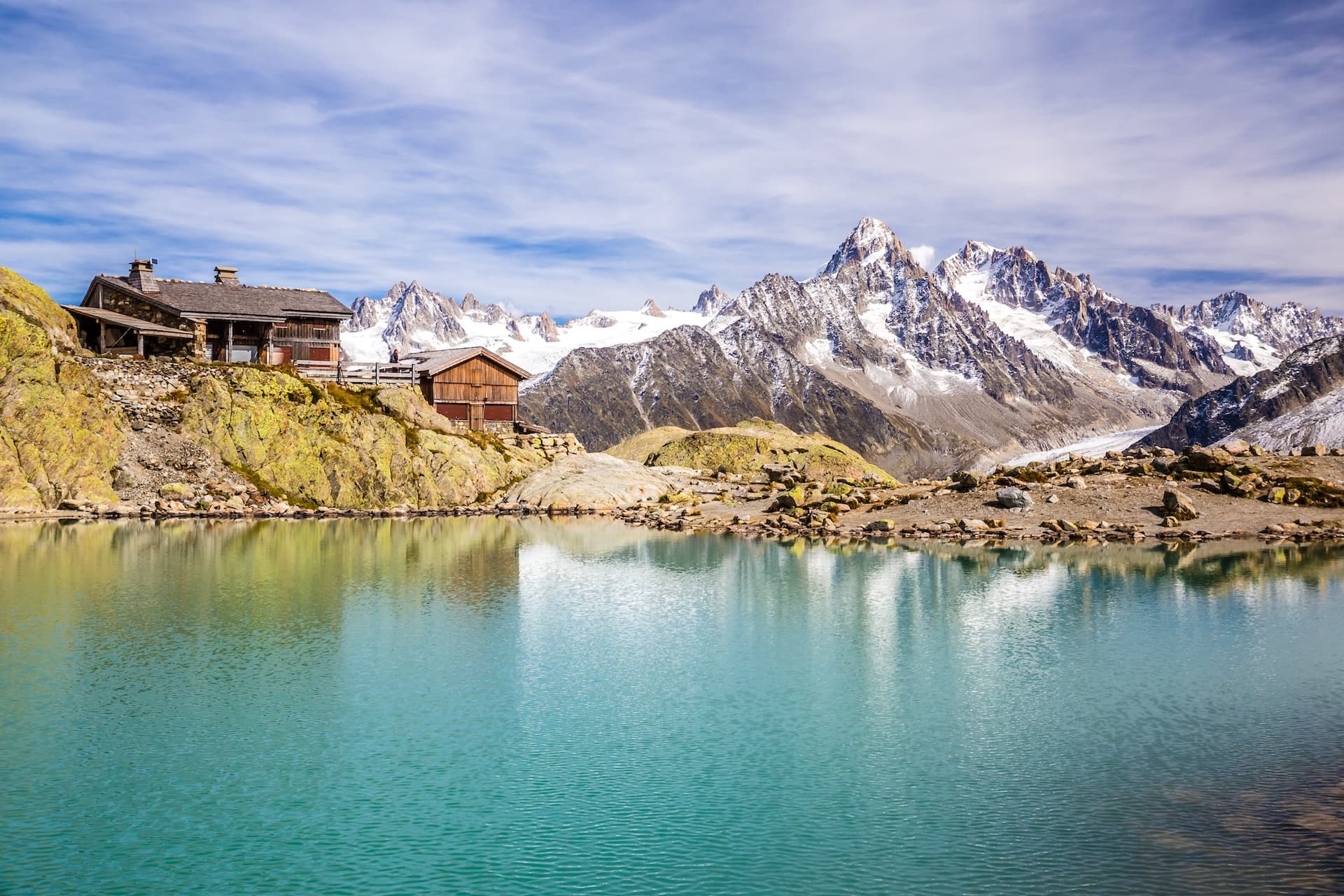 Wooden refuge near turquoise alpine lake with snow-capped mountains reflected