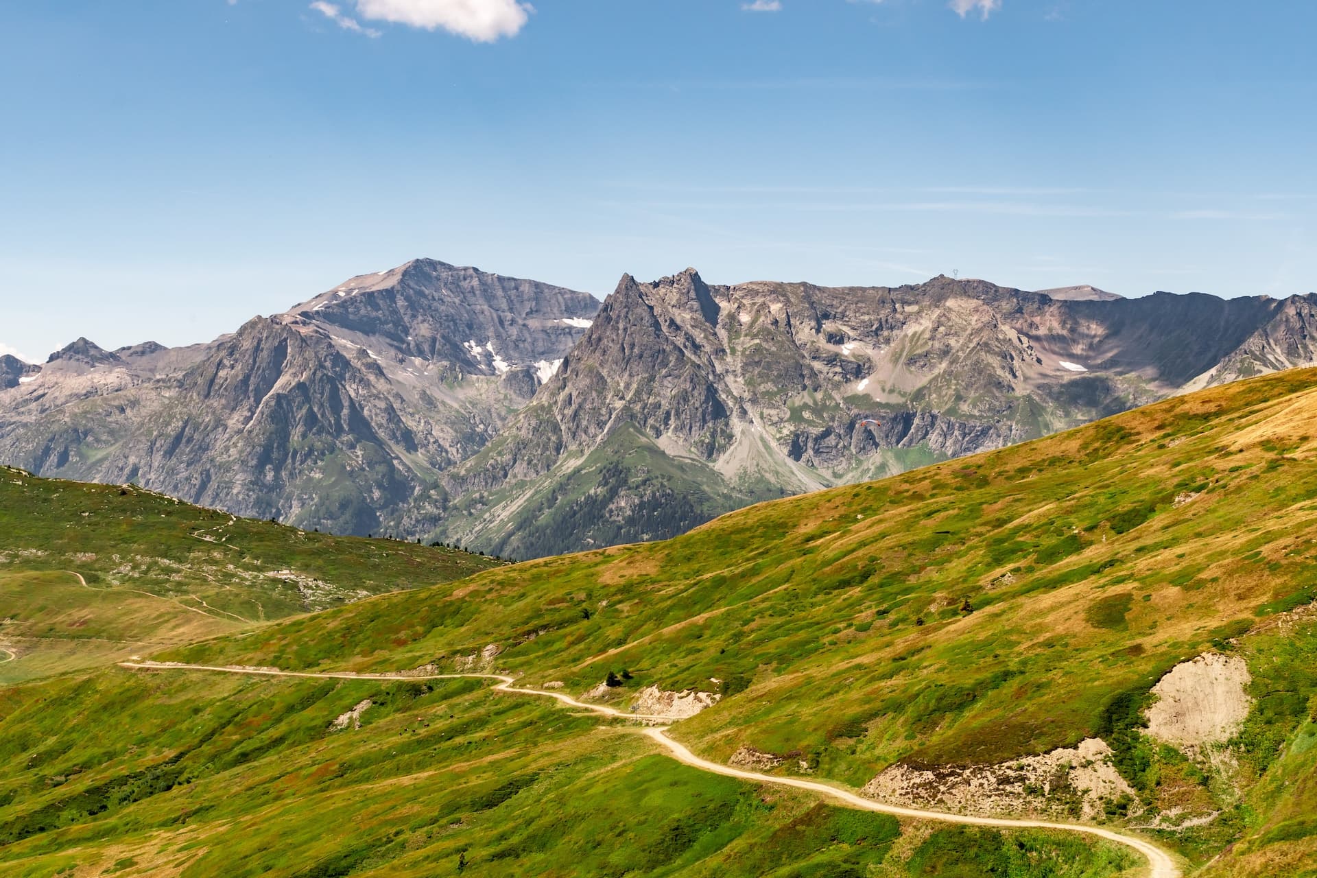 Winding dirt trail on green alpine hillside with rocky mountains and blue sky, Col de Balme.