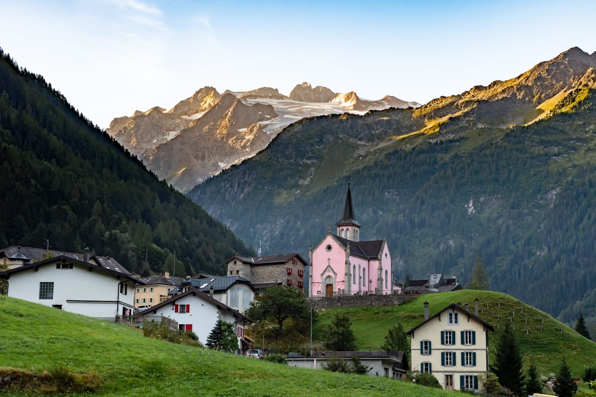 Alpine village with pink church nestled in green valley below sunlit, glaciated mountains.