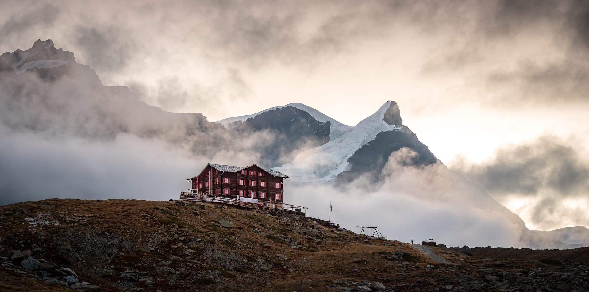 Mountain refuge hut on rocky slope with glacier peaks shrouded in fog at sunrise or sunset.